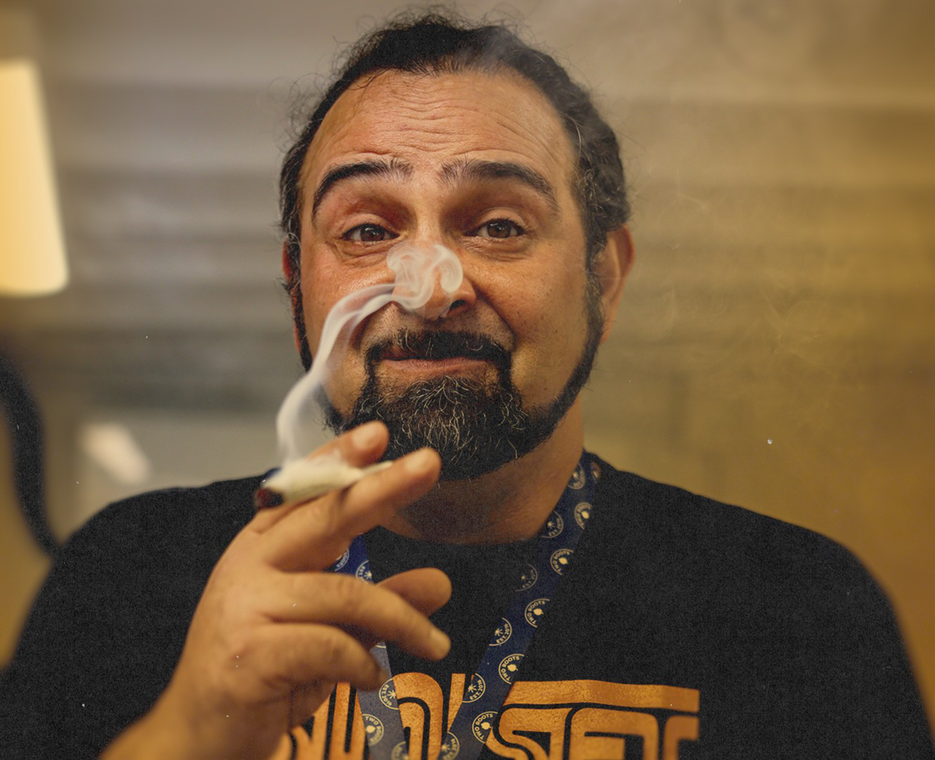 A man with a beard and mustache smokes a rolled cigarette, looking directly at the camera with a relaxed expression and raised eyebrows.