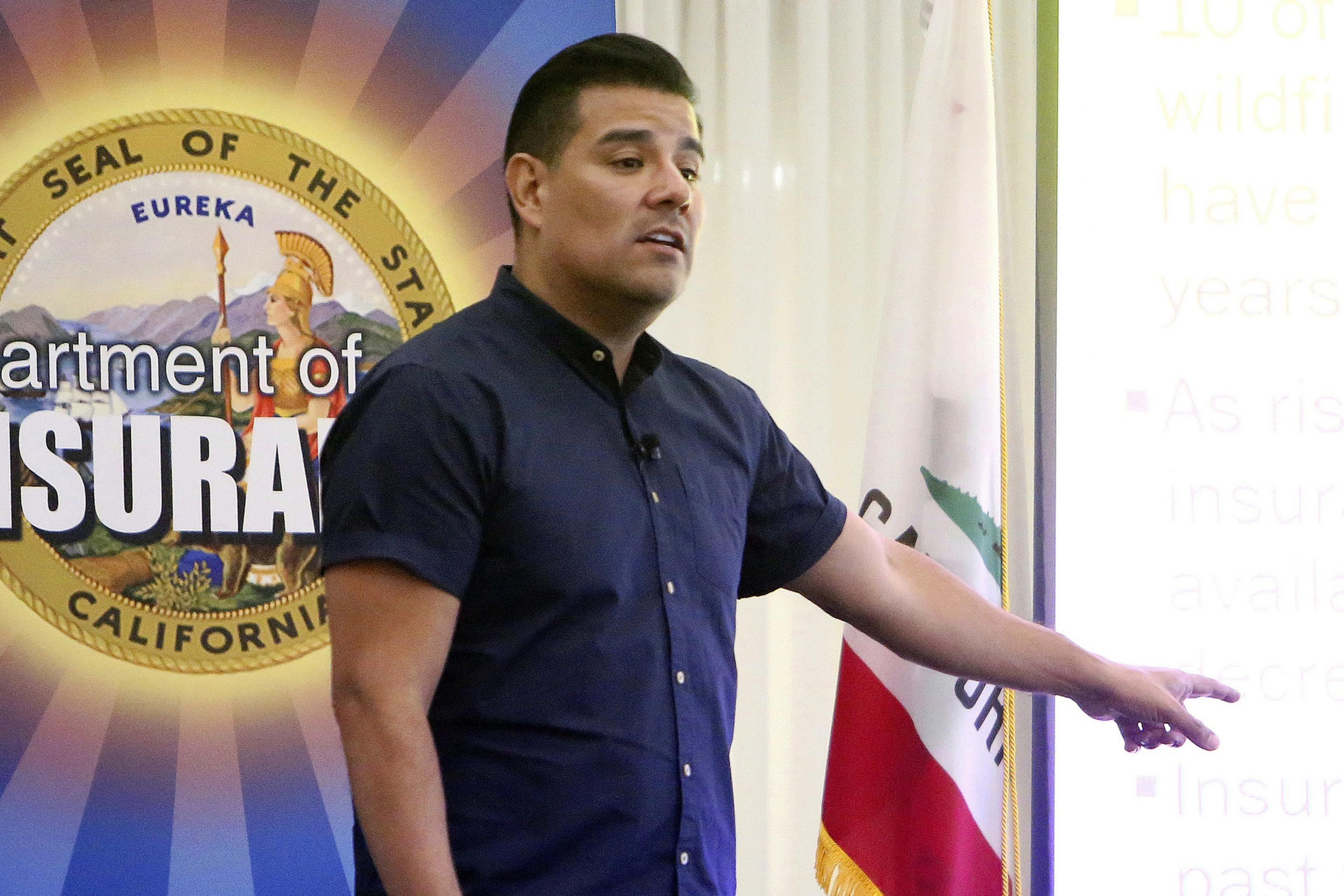 A man speaking in front of a California Department of Insurance seal, pointing with a concerned expression.