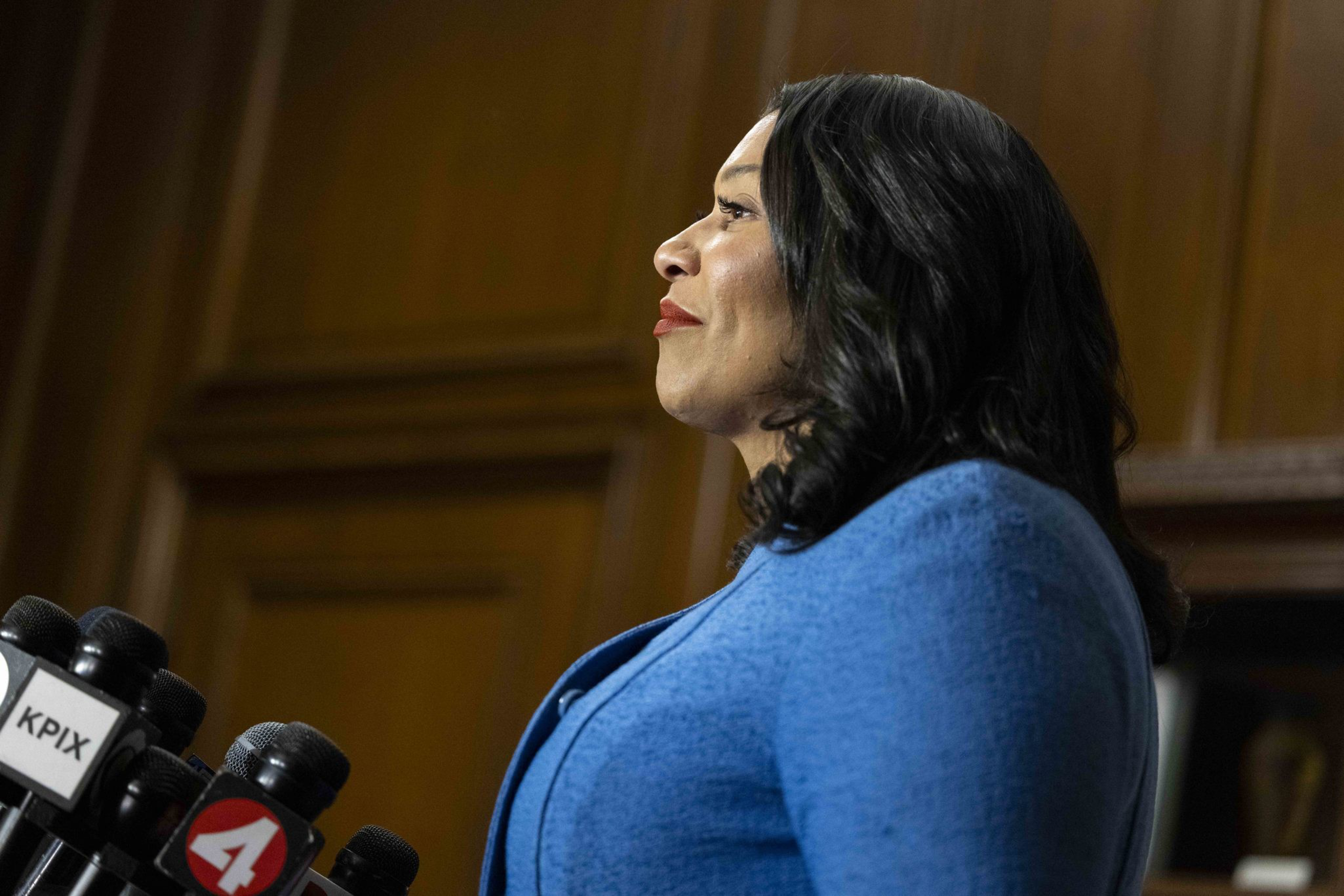 A woman in a blue outfit stands at a podium with multiple microphones. She appears to be speaking or addressing an audience in a formal setting.