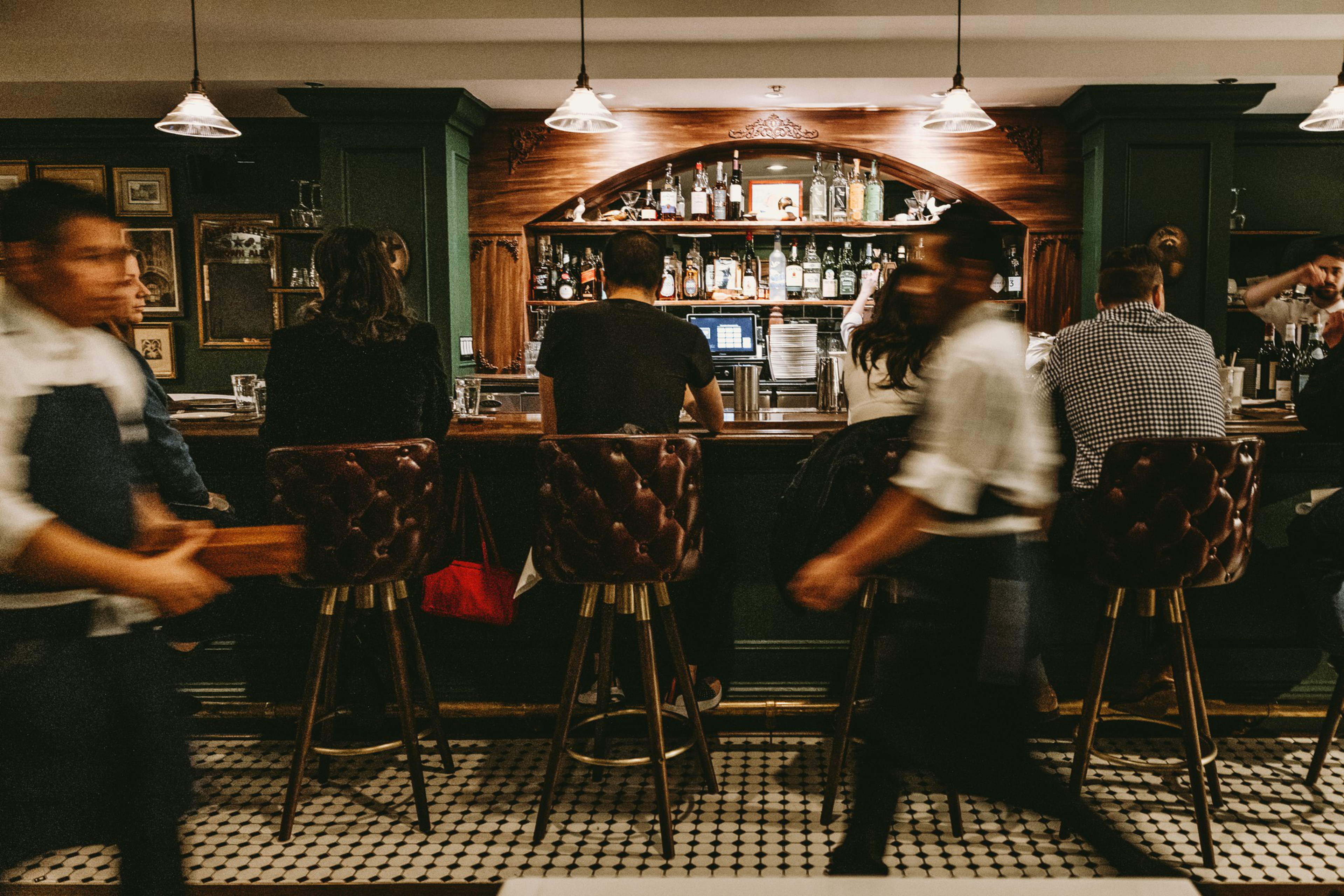 A dimly lit bar with patrons seated on tufted leather stools and two blurred servers moving in the foreground.