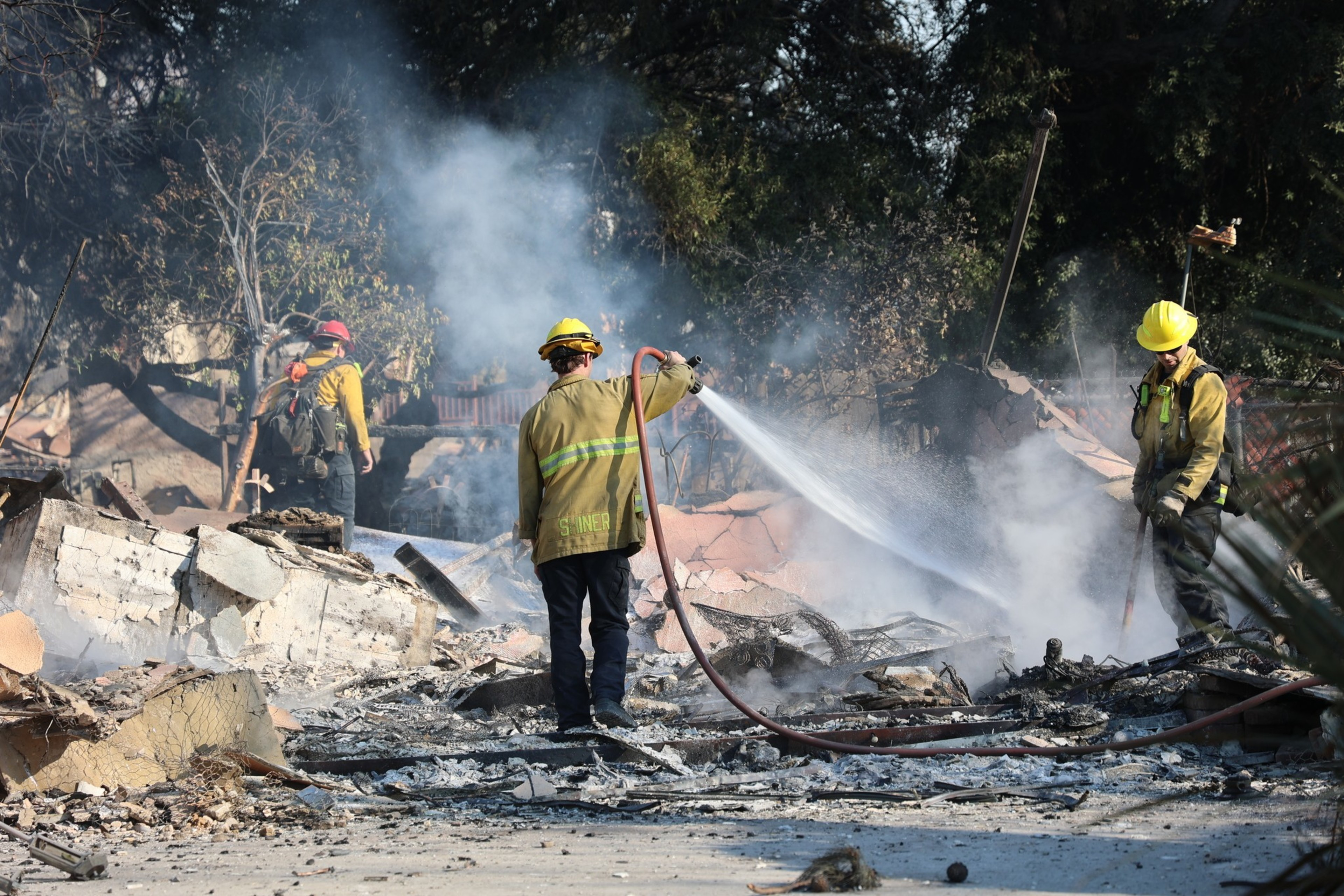 Firefighters in yellow gear spray water on smoldering rubble, surrounded by smoke and debris, amid a scene of destruction with trees in the background.