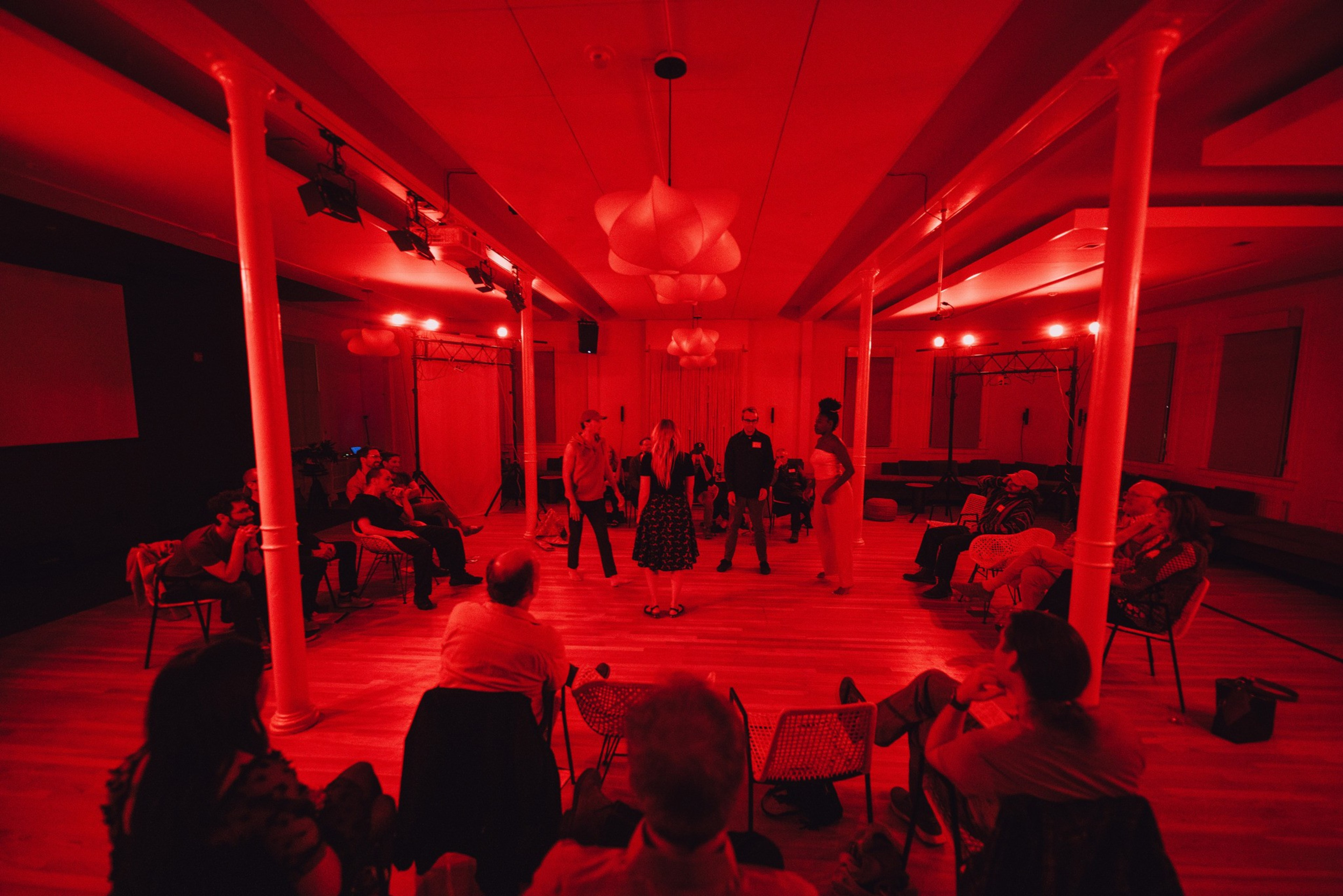 People sit in chairs arranged in a circle around a group of four standing individuals in a dimly lit room bathed in red light.