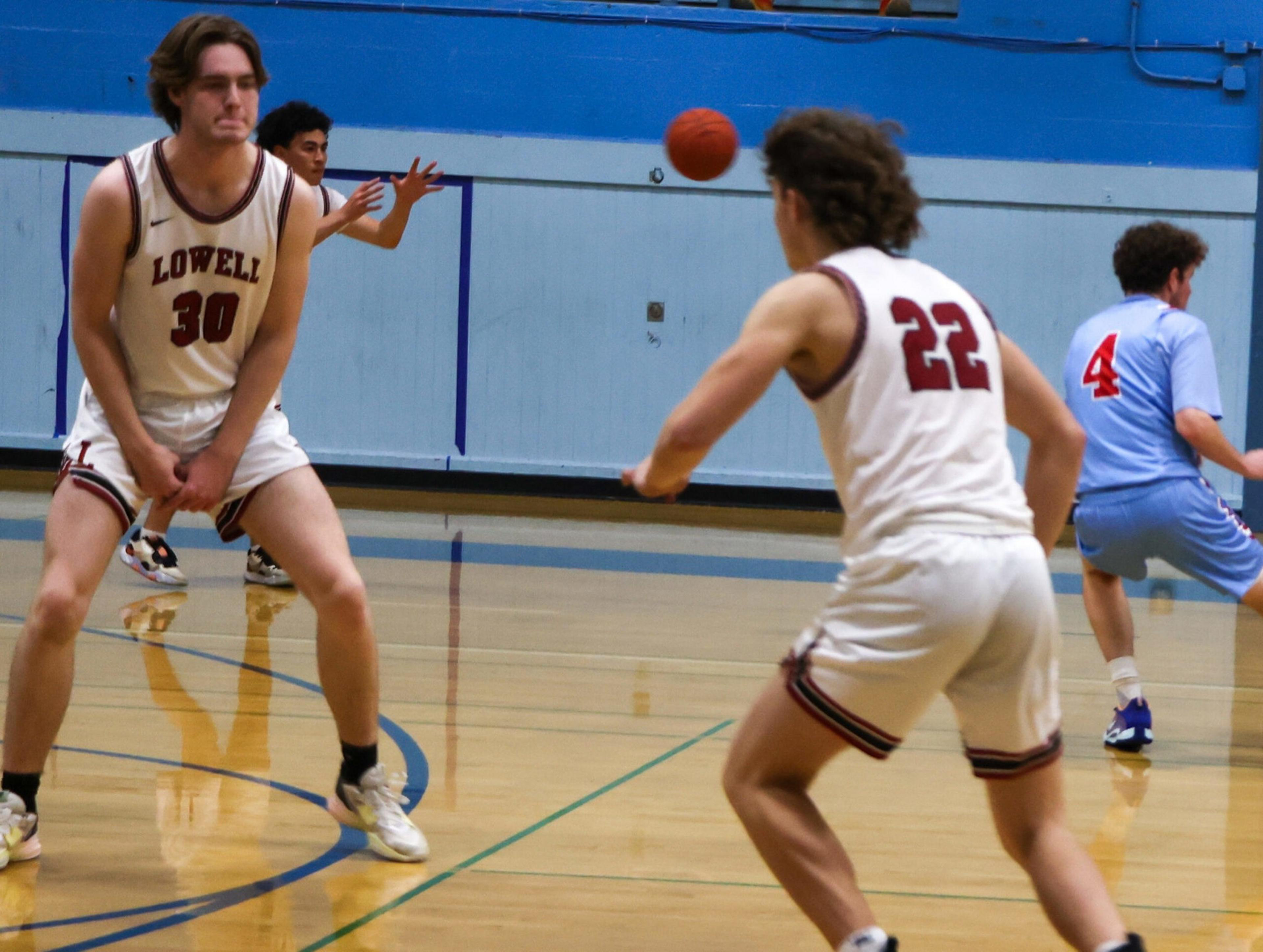 A basketball player wearing a Lowell jersey (number 30) braces to catch the ball being passed by teammate number 22, while opponents in blue watch.