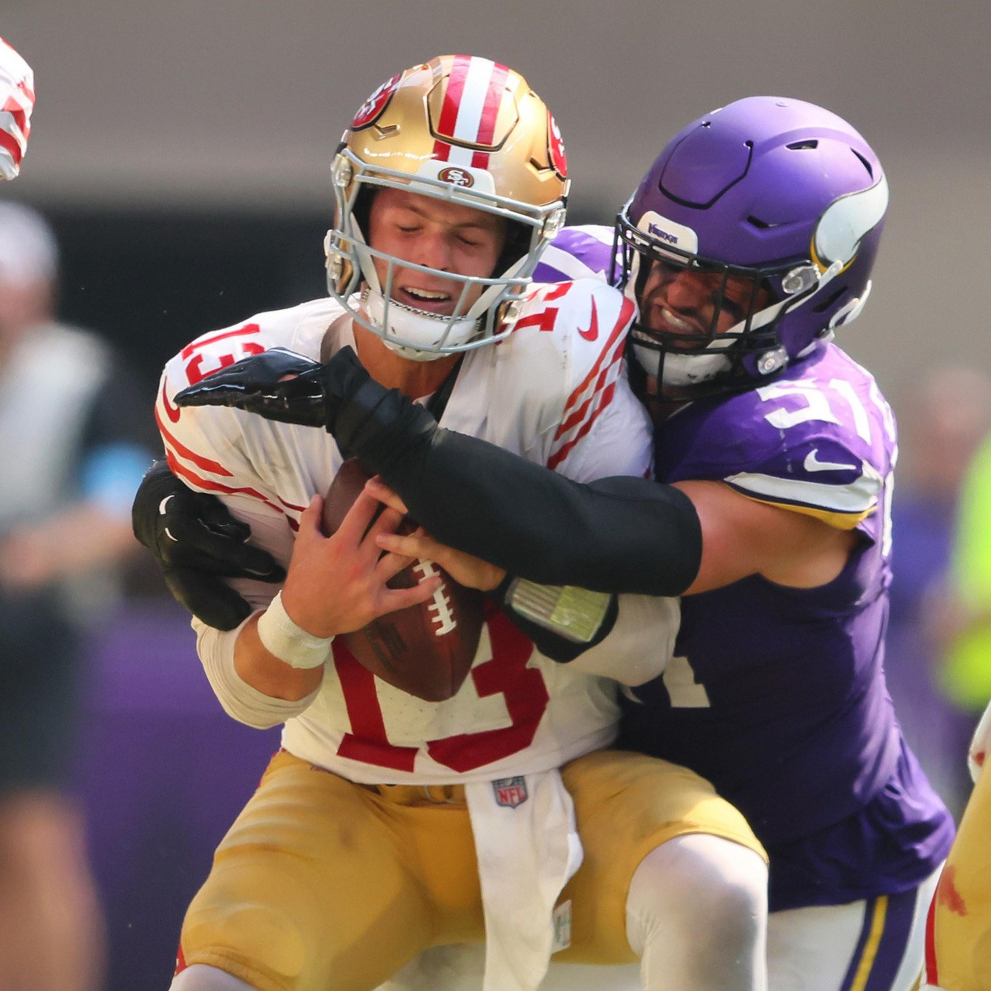 A football player in a purple uniform tackles a player in a white and red uniform holding a football. The scene is intense, with other players around them.