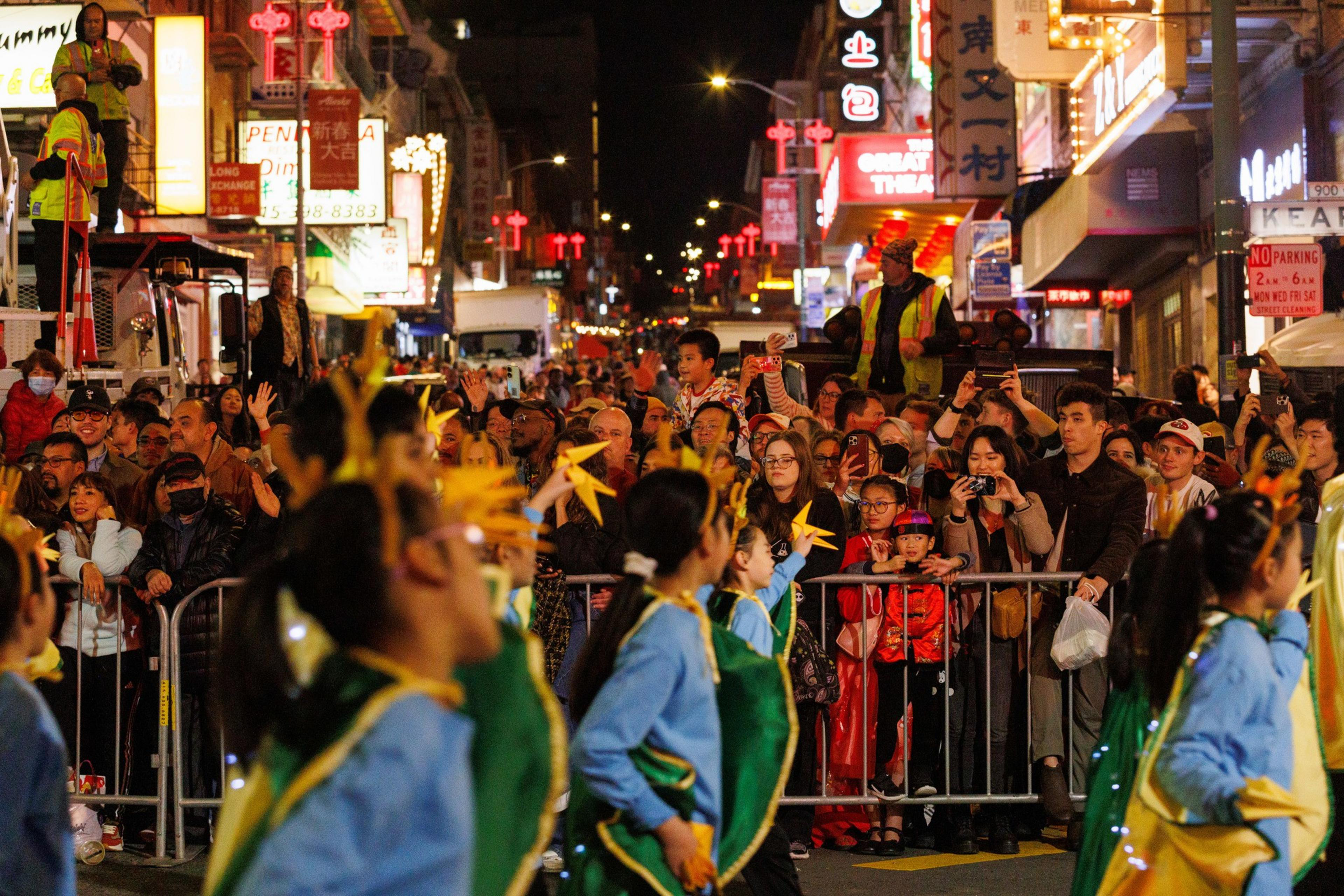 A vibrant street parade at night with dancers wearing colorful costumes. A crowd watches, taking photos, as bright neon signs illuminate the scene.