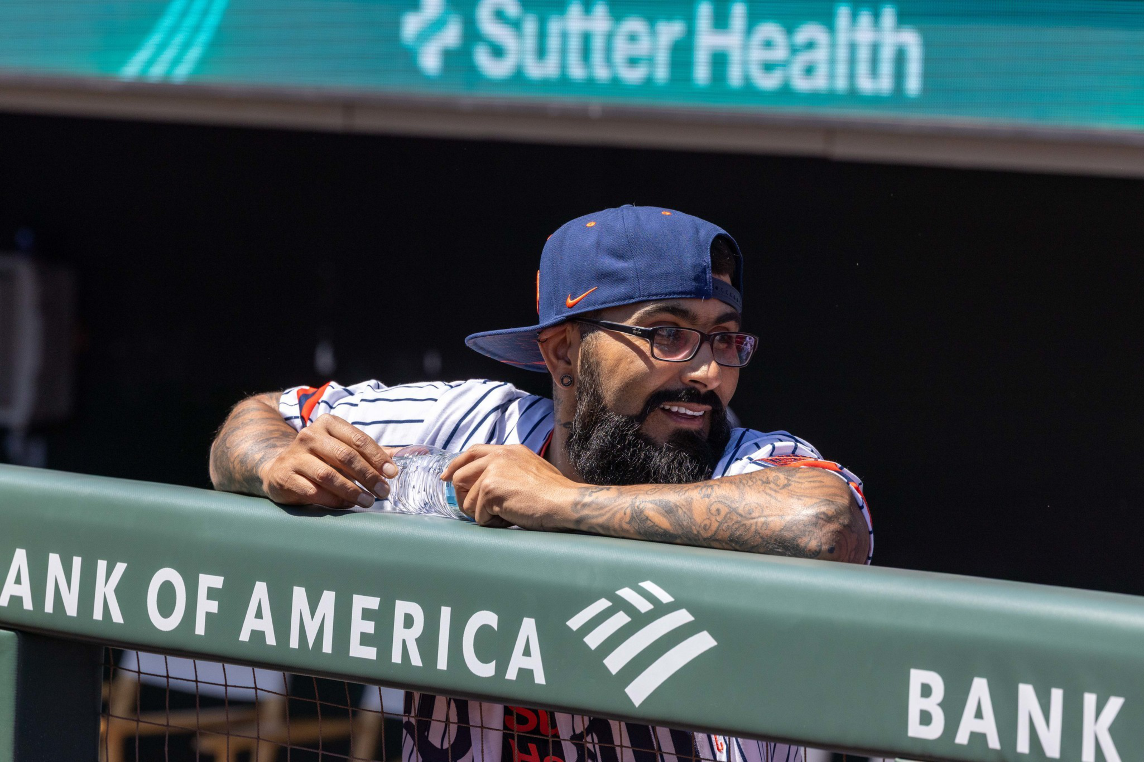 A man with a beard, glasses, and tattoos leans on a railing marked "Bank of America," wearing a baseball cap backwards and a sports jersey.