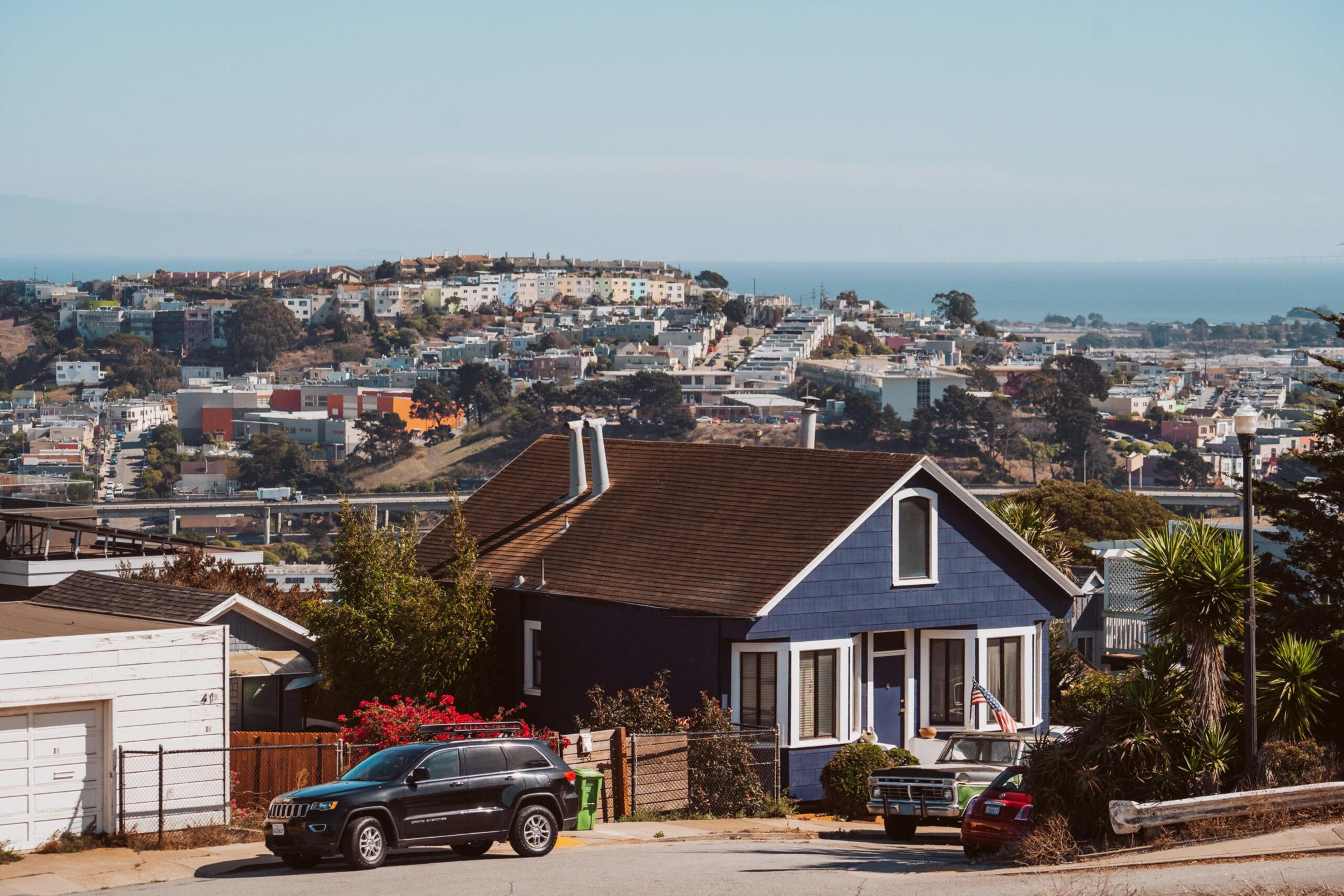A dark blue house with white trim sits on a hill, surrounded by cars, trees, and a view of a dense cityscape reaching toward a distant ocean.