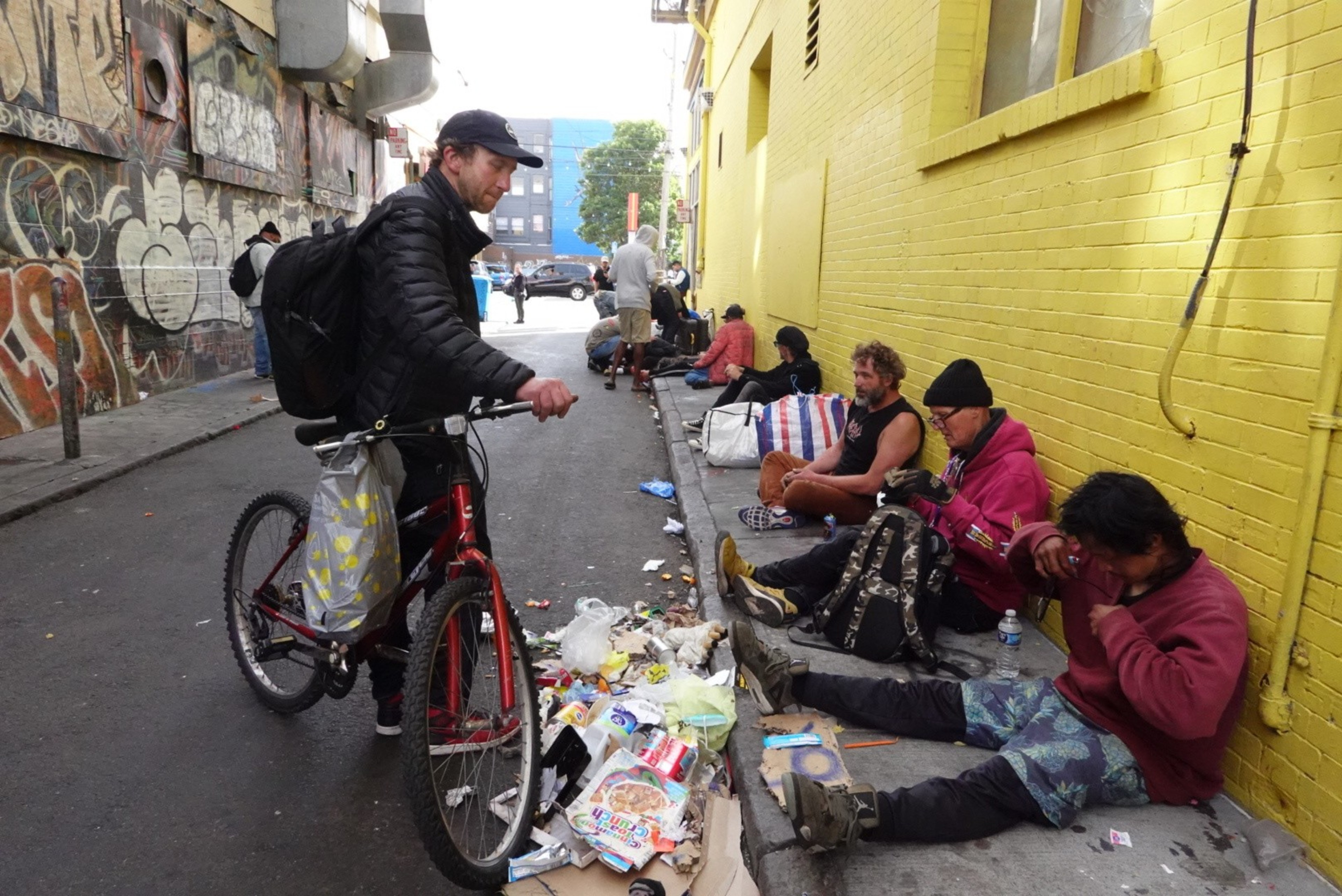 A man with a bike observes several people sitting along a yellow brick wall in an alley. There is scattered trash on the ground, and graffiti covers the opposite wall.