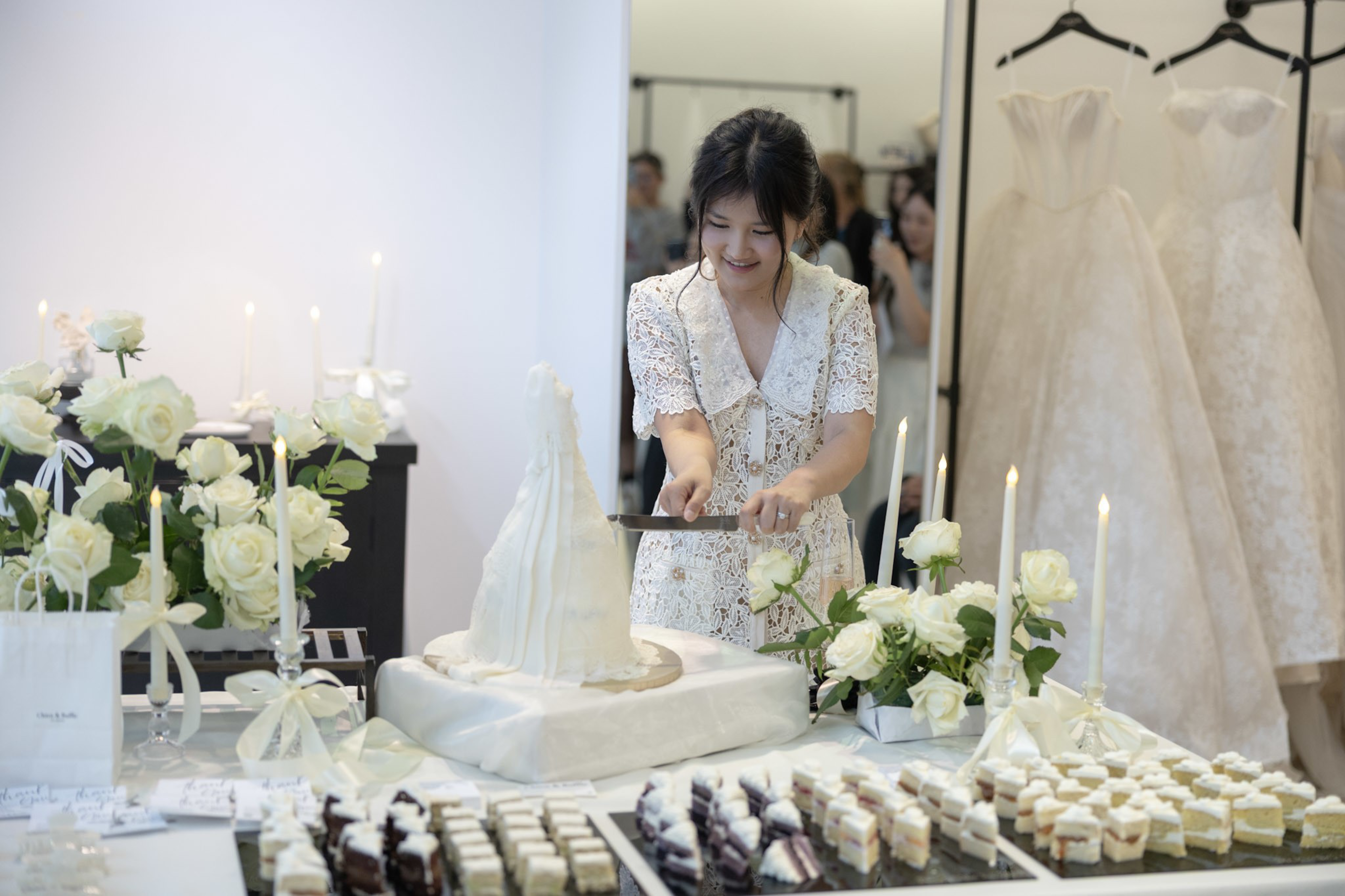 A young woman wearing a white lace dress is smiling as she cuts a white cake shaped like a wedding dress, surrounded by white roses and desserts.