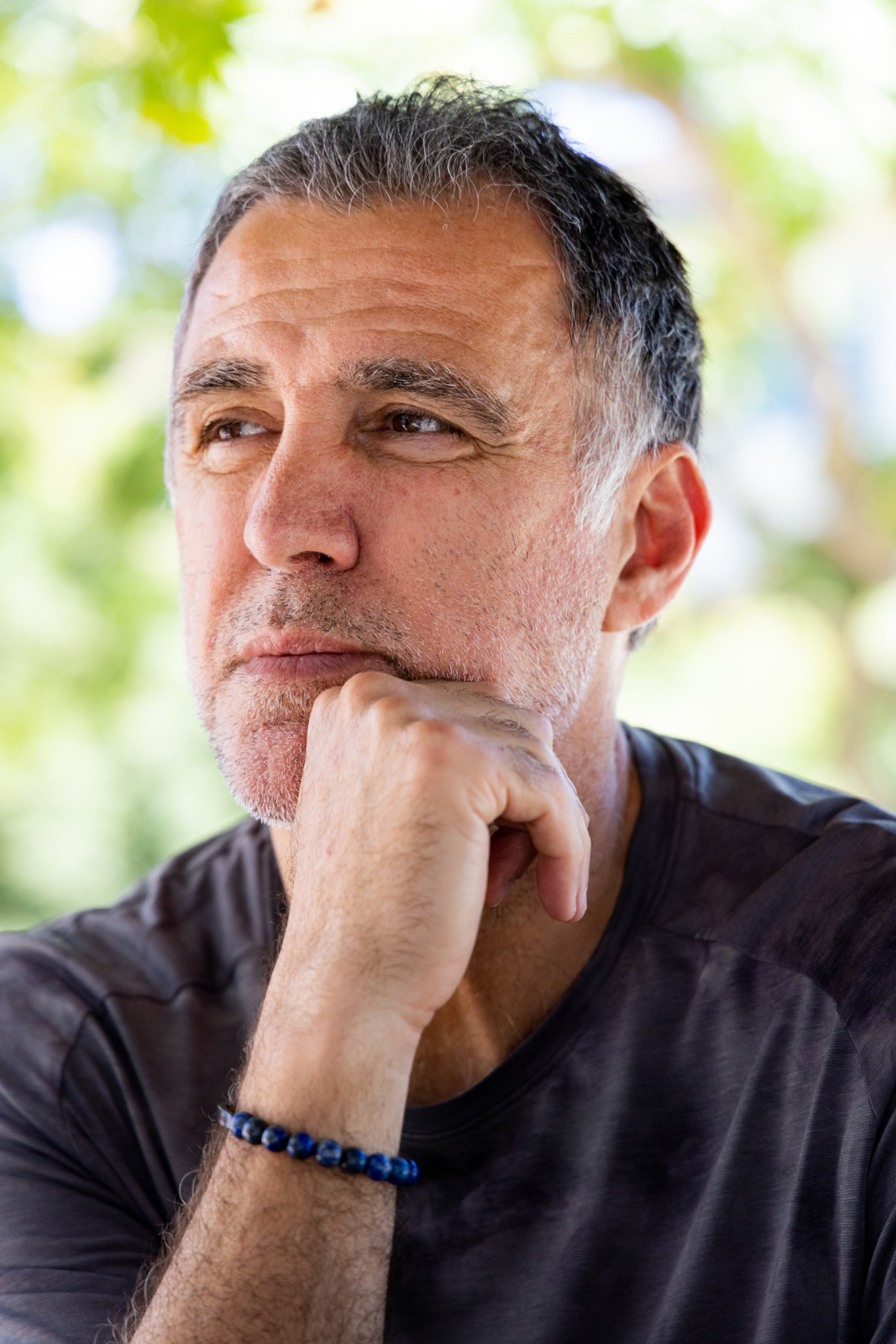 A middle-aged man with salt-and-pepper hair rests his chin on his hand, wearing a dark shirt and a blue beaded bracelet, looking thoughtfully to the side.