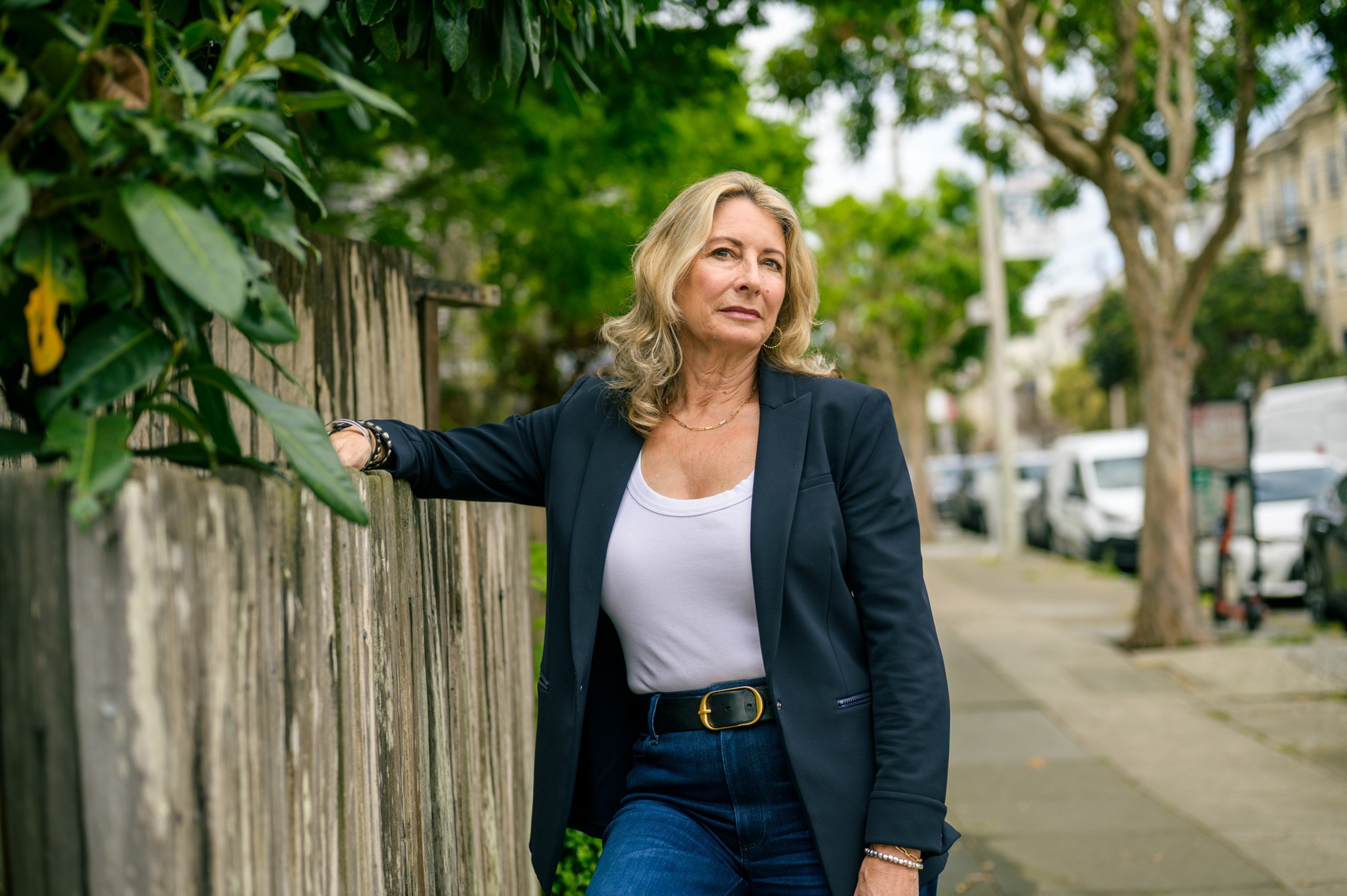 A woman with blonde hair wearing a white top, black blazer, and blue jeans leans on a wooden fence on a tree-lined sidewalk.