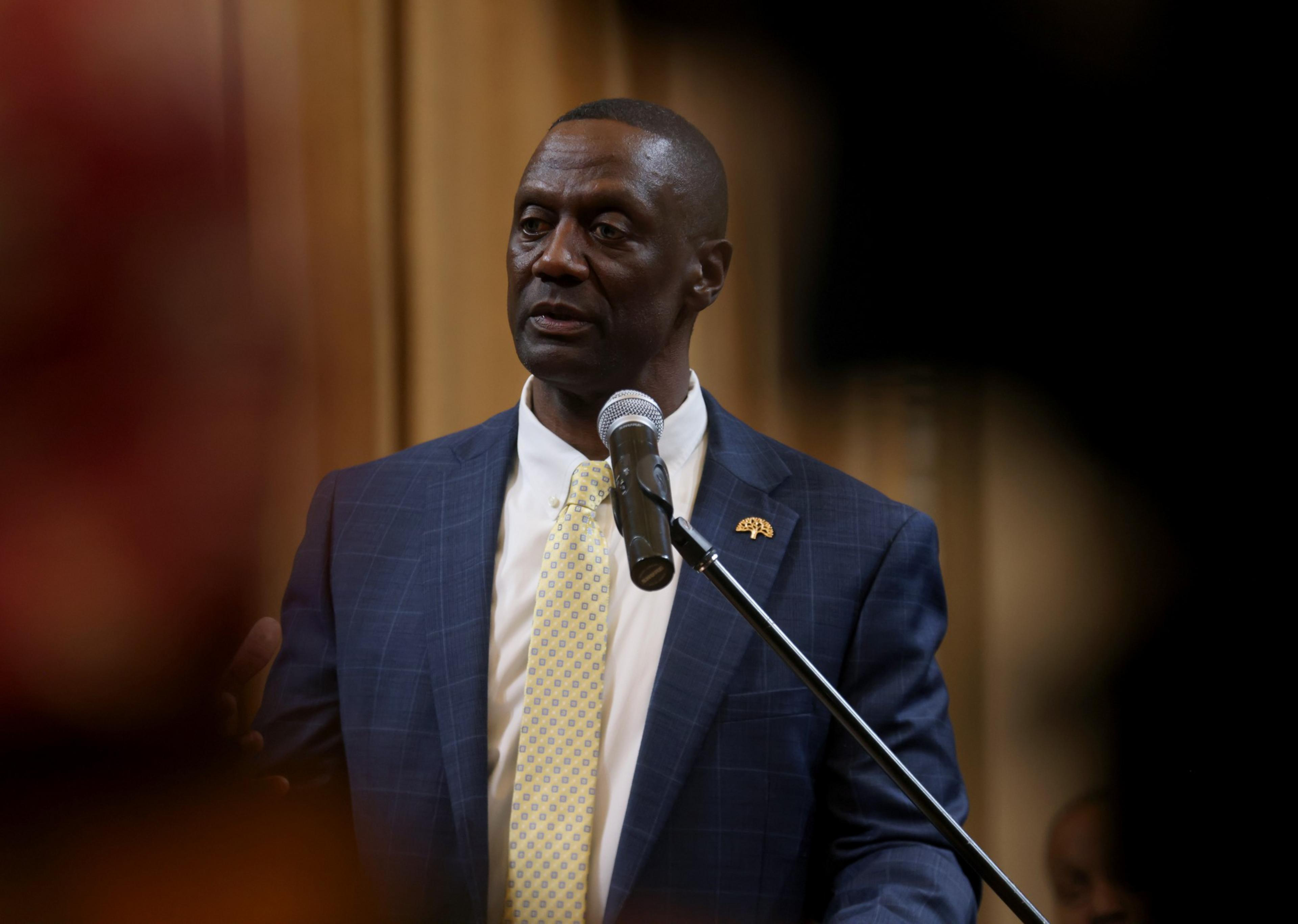 A man in a blue suit and yellow tie speaks into a microphone with a serious expression against a blurred indoor background.