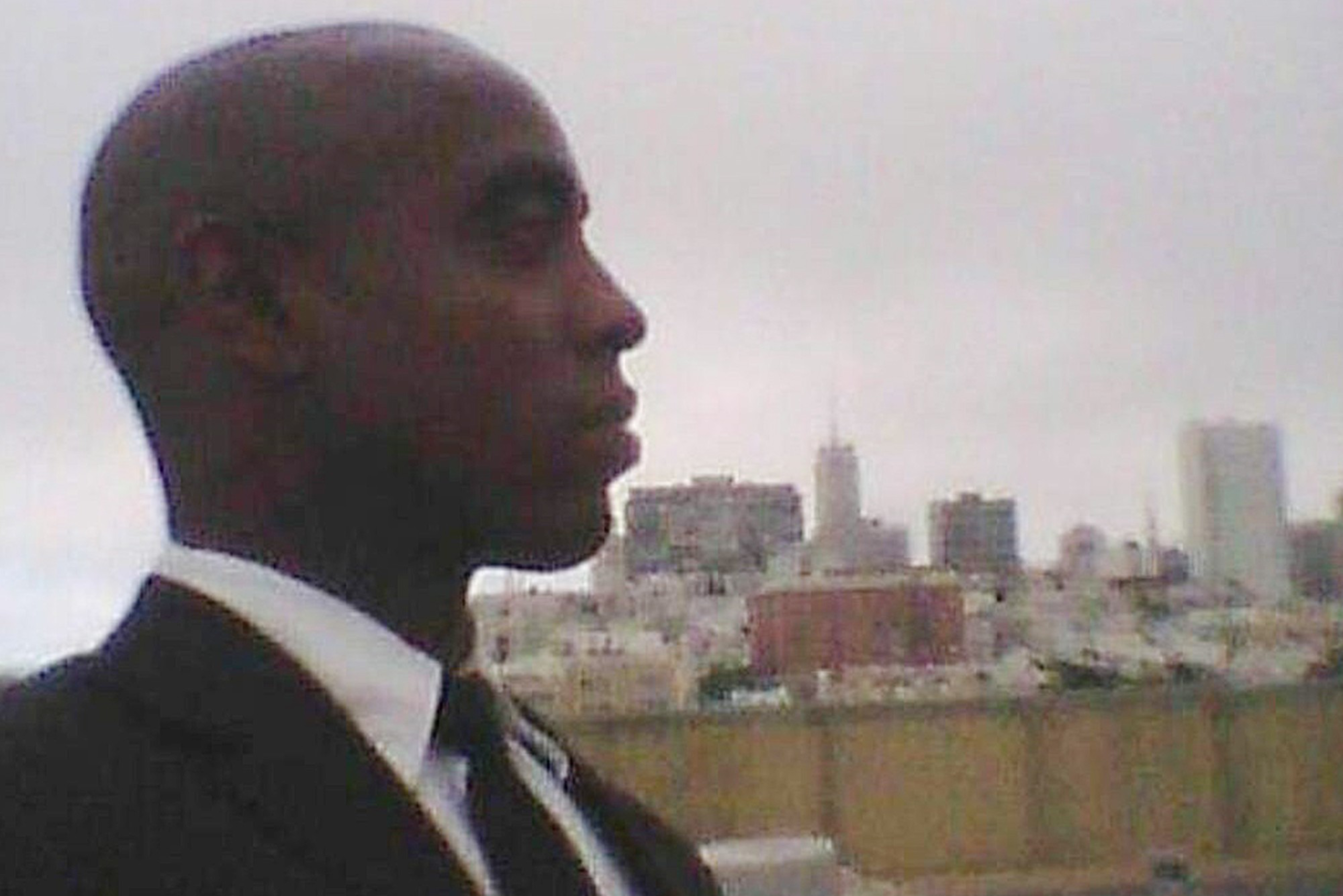A man in a suit and tie faces left with a city skyline blurred in the background under a gray, overcast sky.