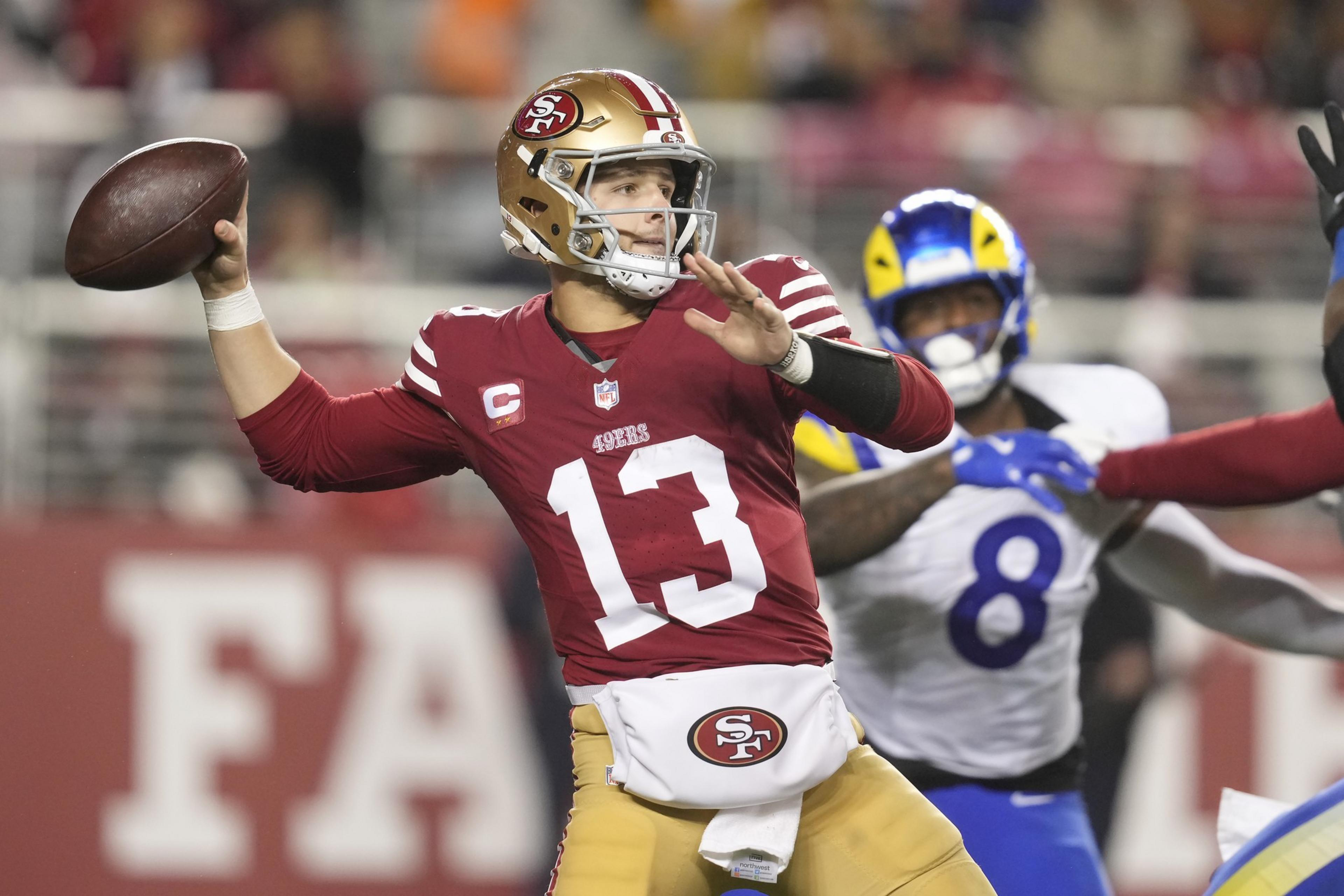 A football player in a red jersey and gold helmet is poised to throw a ball. He's wearing number 13 and is surrounded by other players on a field.