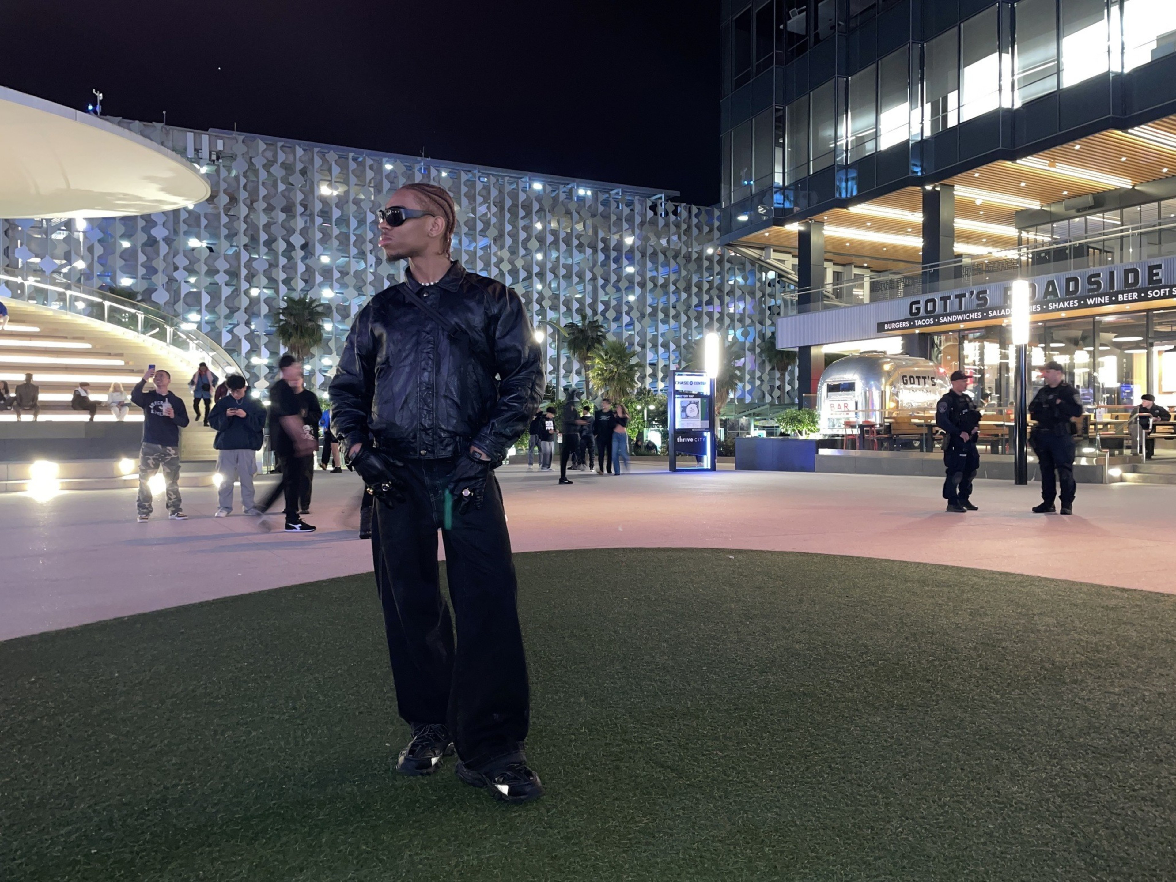A person stands confidently on artificial turf at dusk, with a patterned building and a restaurant behind them.