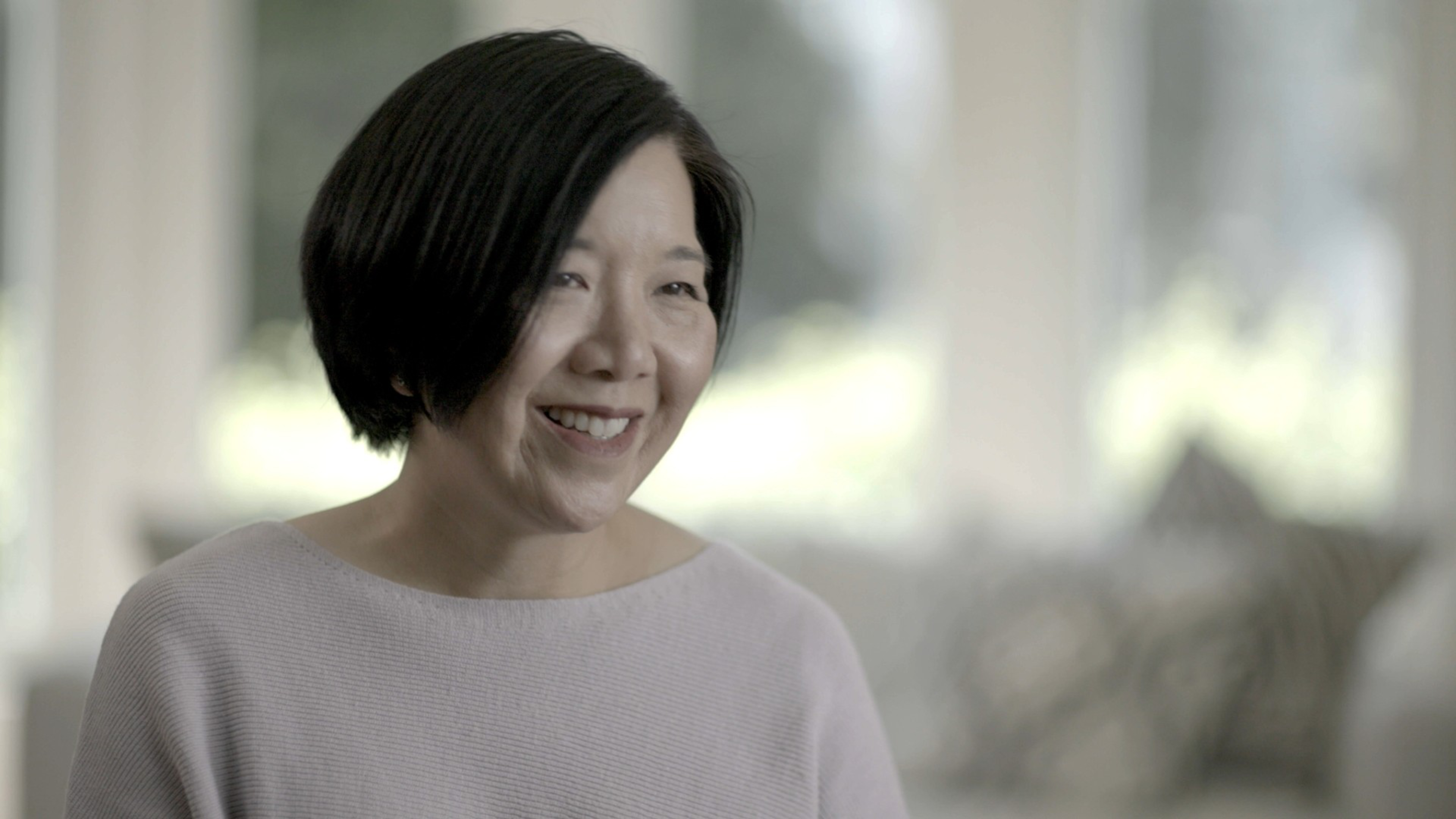 A smiling woman with short black hair wears a light purple sweater, sitting indoors with soft natural light and a blurred background.