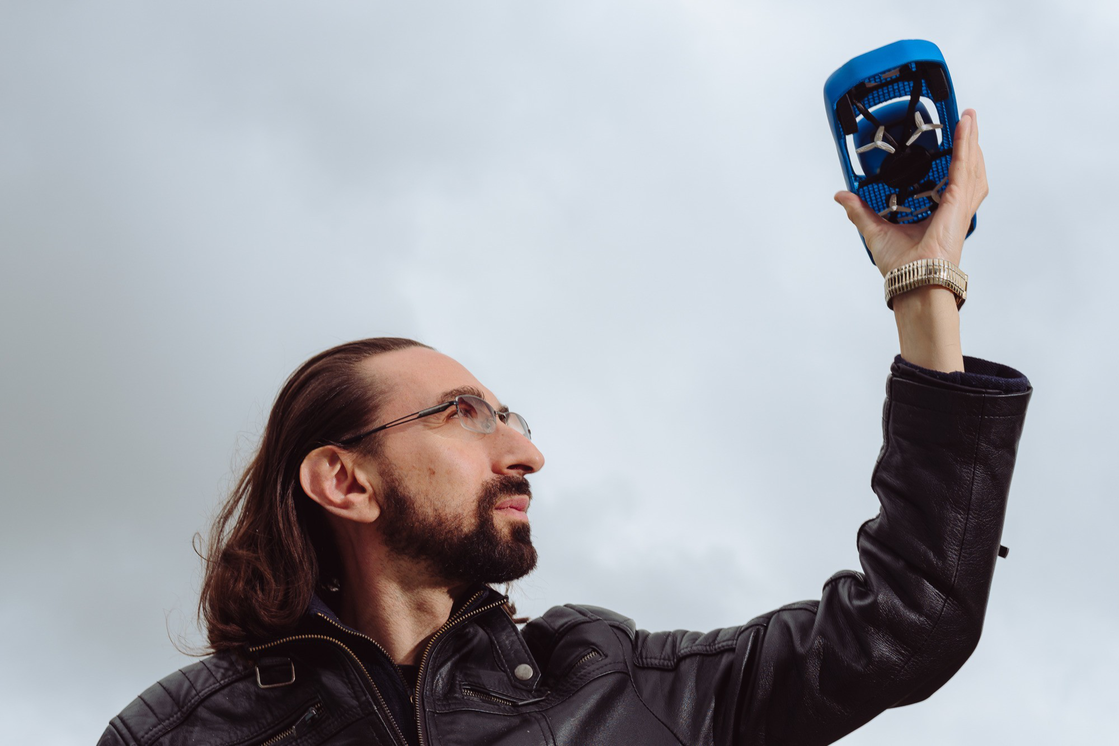 A man with long hair and a beard, wearing glasses and a leather jacket, holds a small, blue drone up to the sky. The background is overcast.