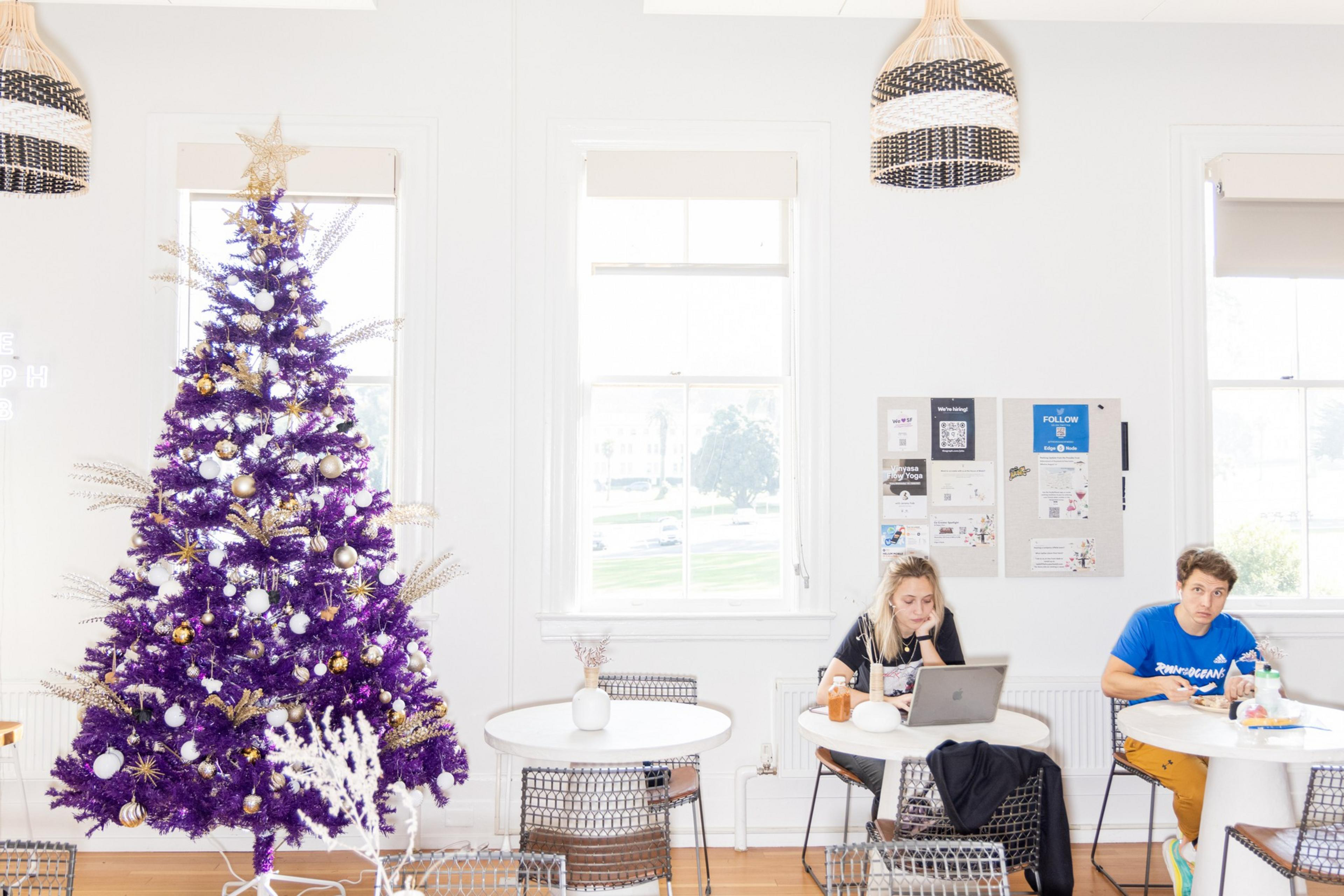 A vibrant purple Christmas tree adorned with gold and white decorations stands beside two people in a bright room, one on a laptop and the other eating.