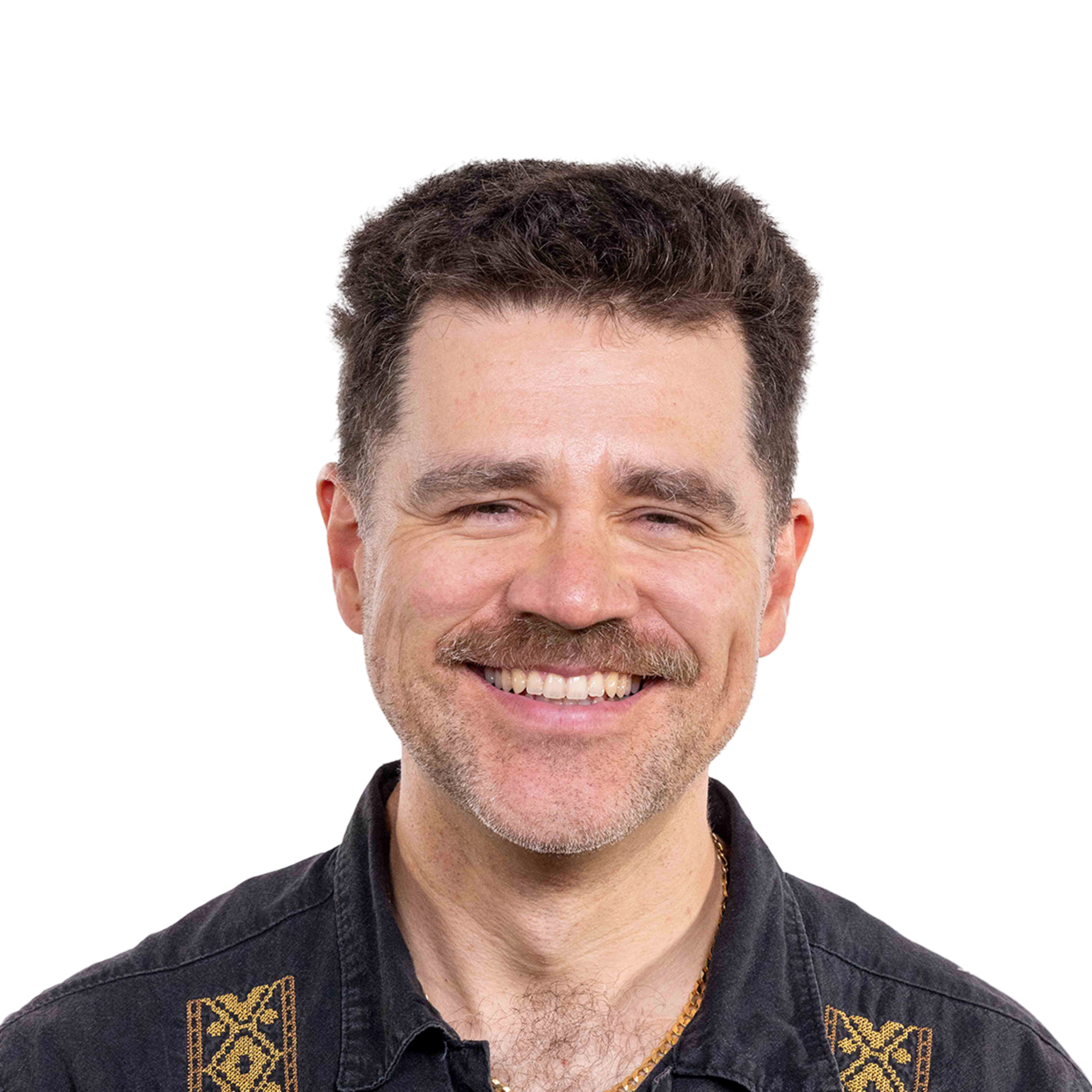 A smiling man with dark curly hair and a mustache wears a black shirt with gold embroidered patterns and a gold chain necklace.