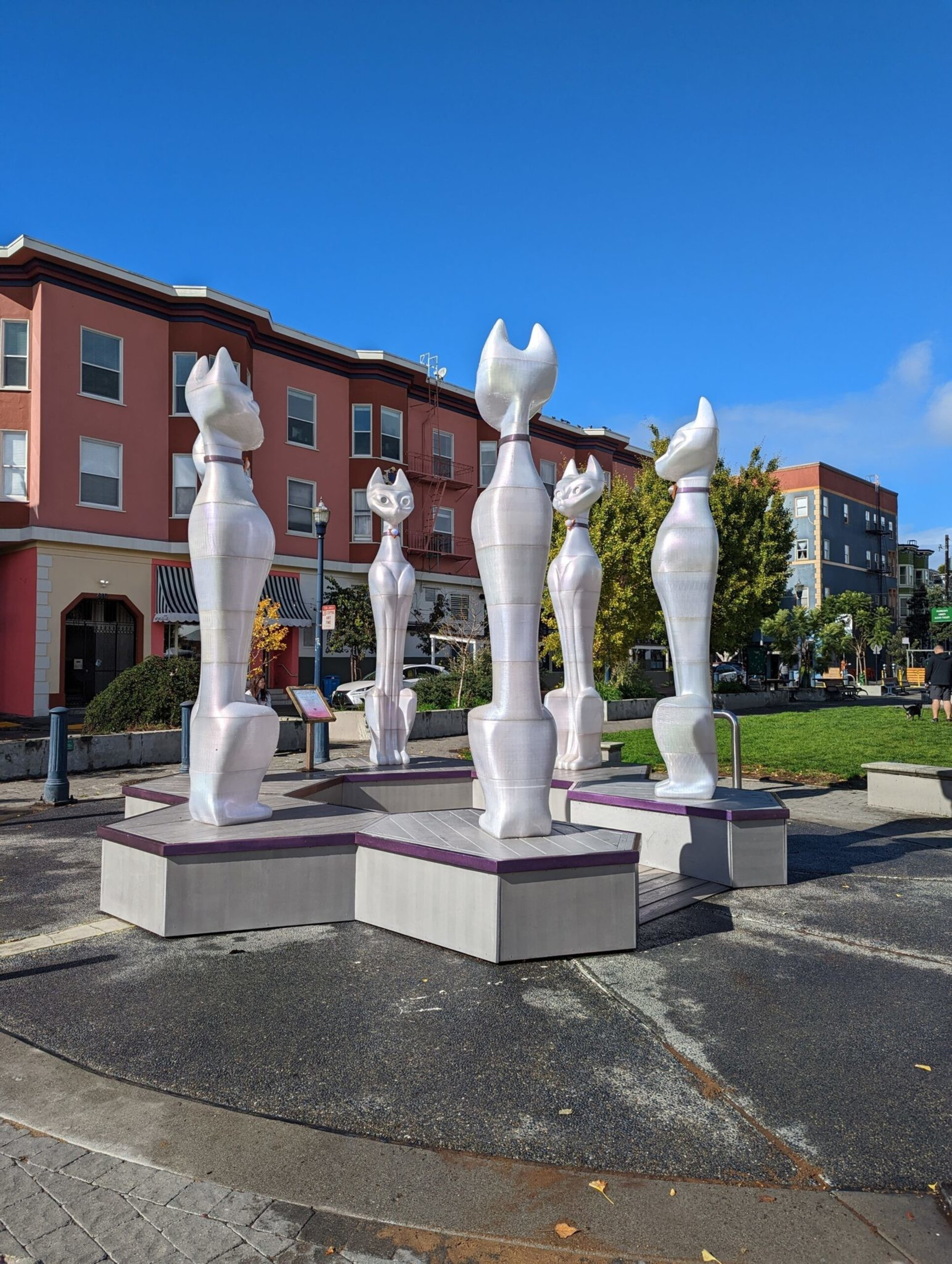 Five tall, white cat sculptures stand on a multi-level hexagonal platform in a sunny outdoor plaza with buildings and trees nearby.