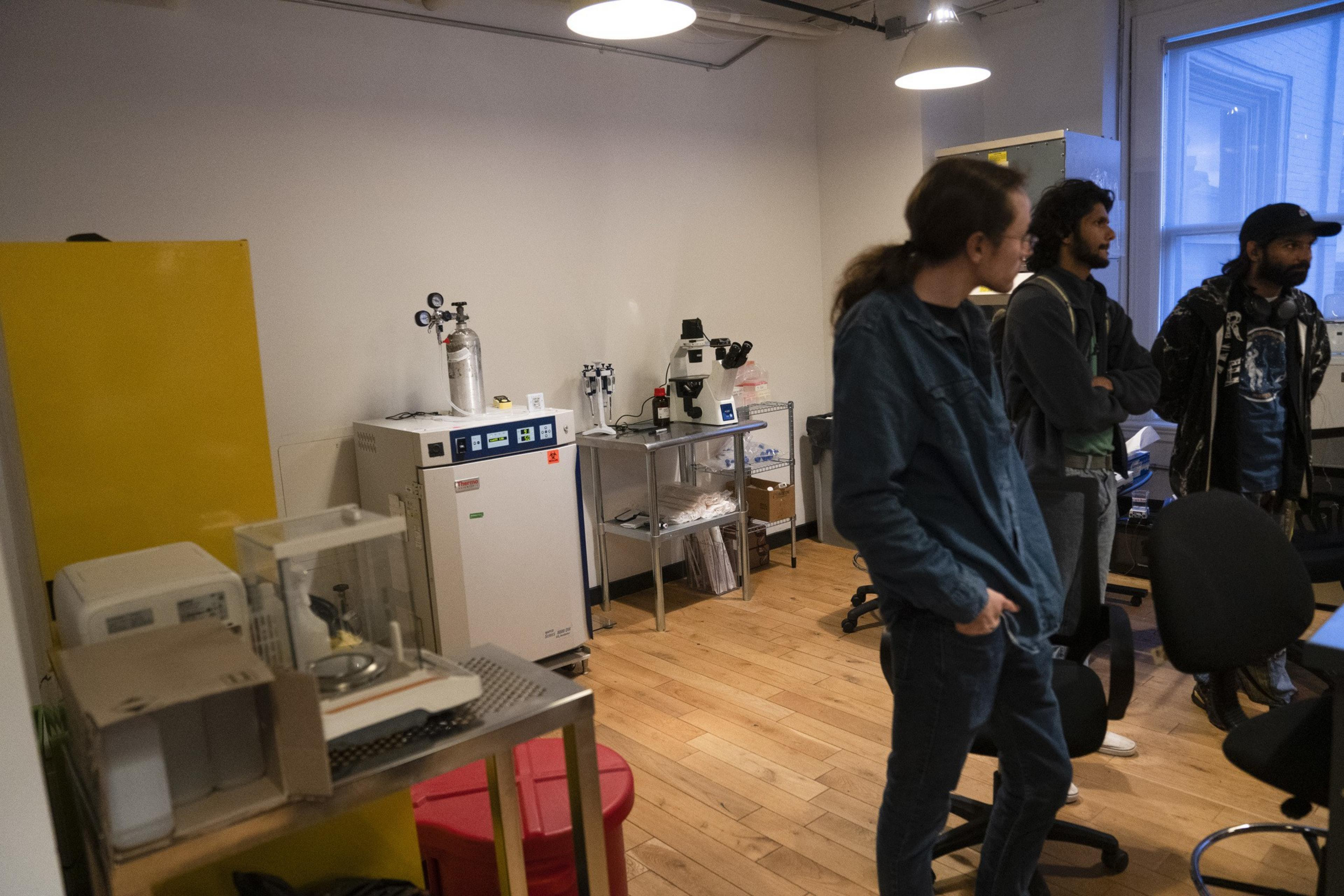 A group of people stands in a lab with a microscope, equipment, and wooden flooring. There's a yellow panel on the left and a window on the right.