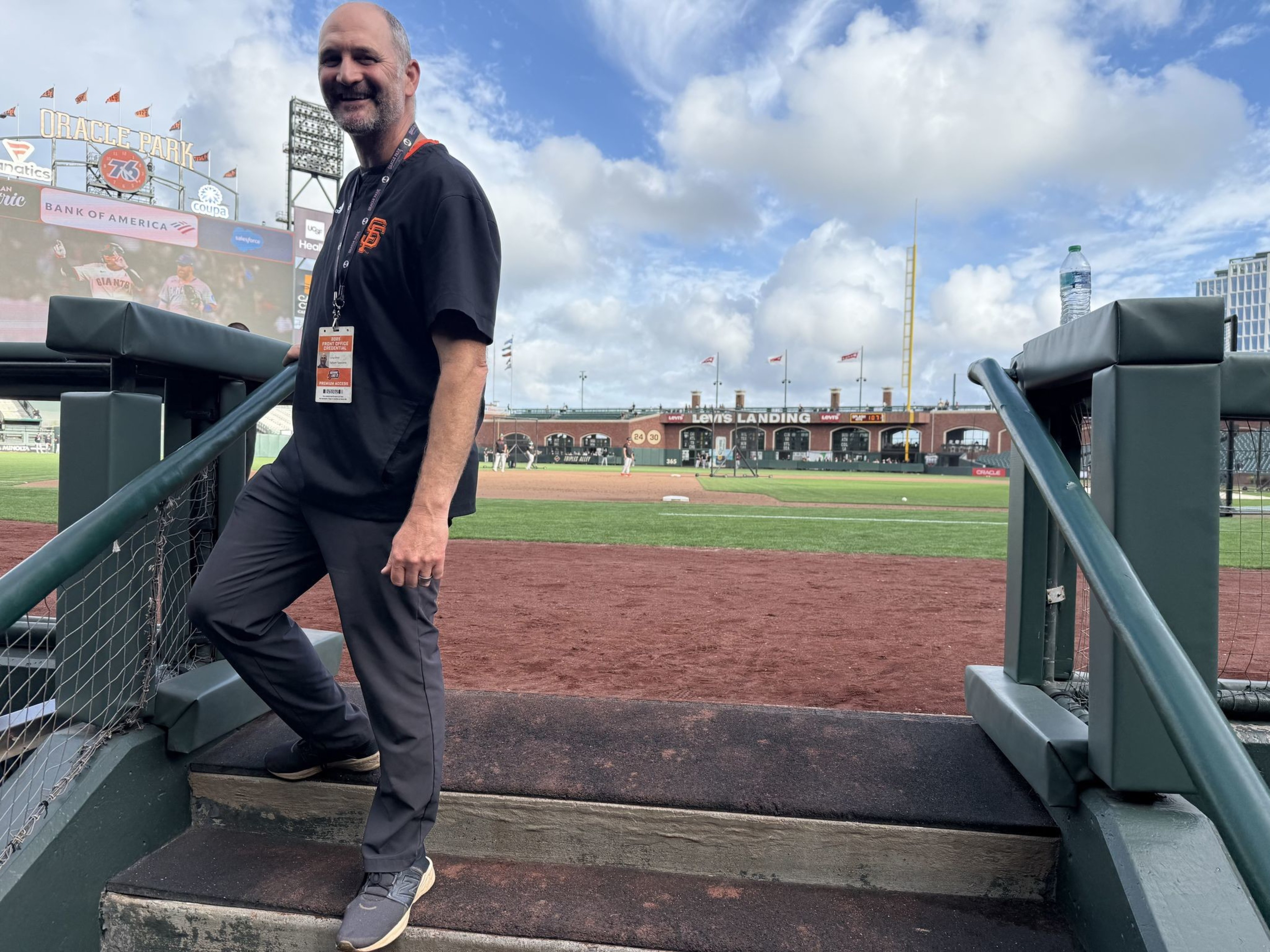 A man wearing a black Giants shirt and badge stands on steps leading to a baseball field at Oracle Park, with the stadium and scoreboard visible behind him.
