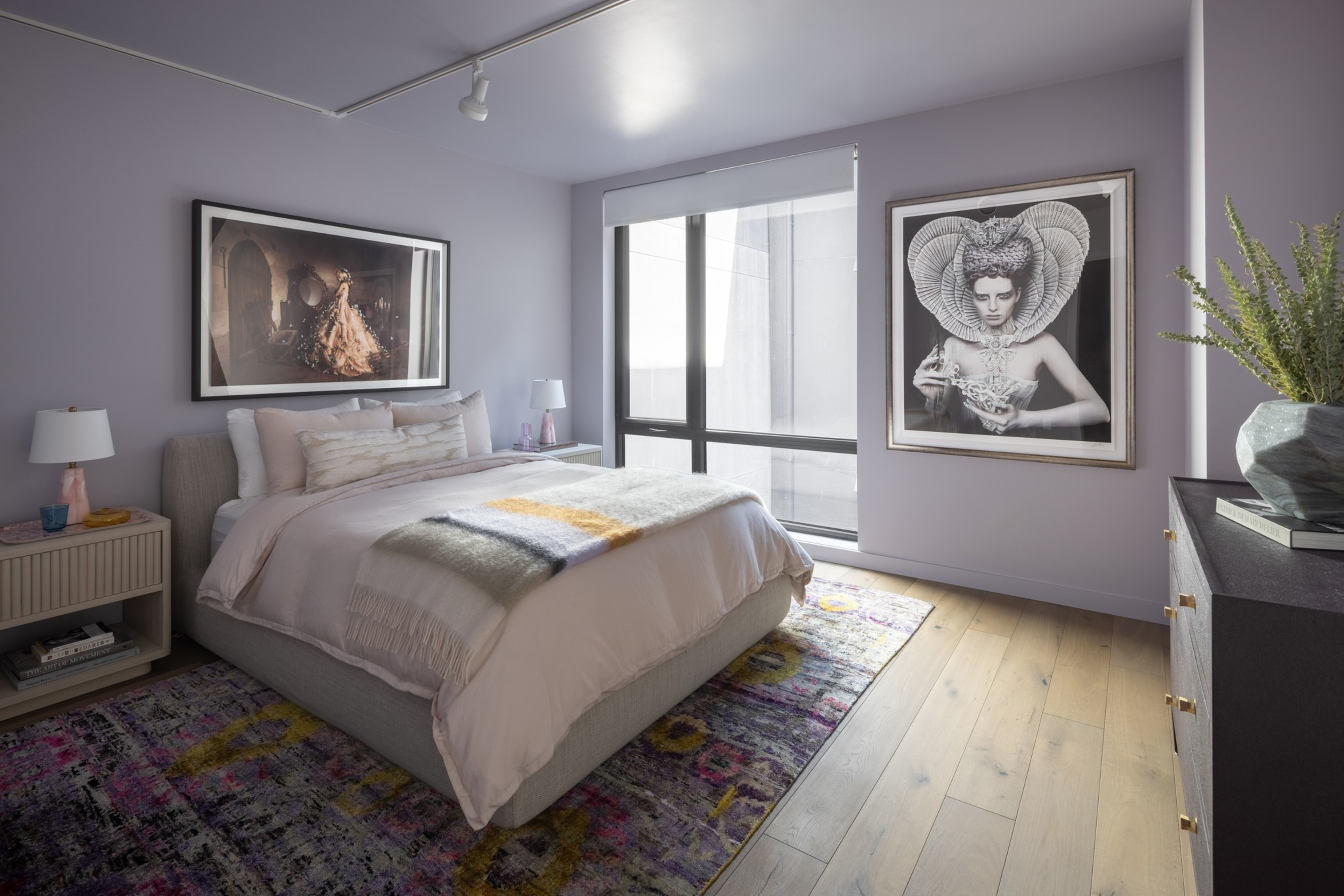 A modern bedroom with a gray bed, soft white bedding, two framed artistic portraits, a colorful rug, wooden floor, and a large window with natural light.