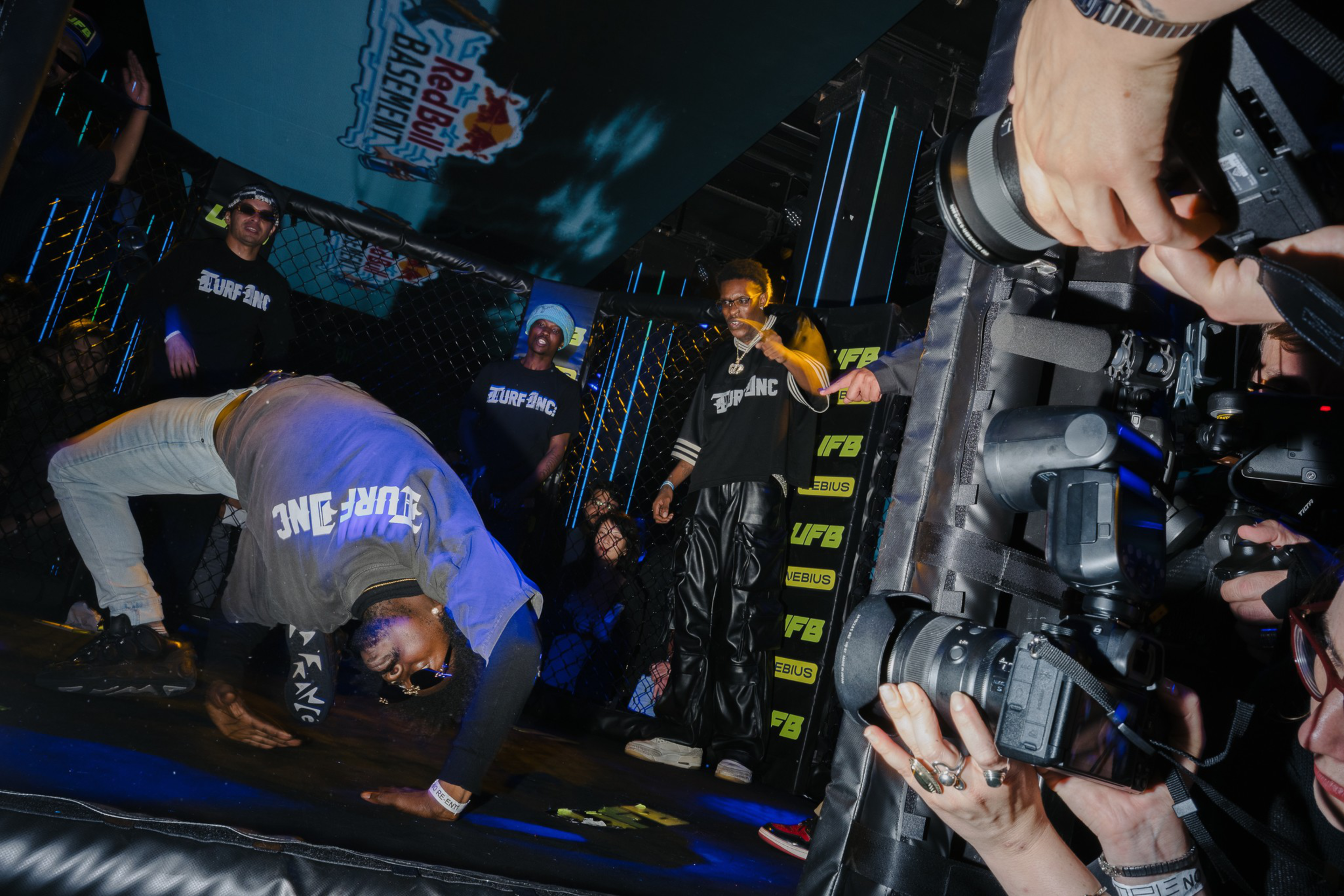 A dancer bends backward on the floor while three people watch, two wearing matching black “SURF ANC” shirts, and photographers capture the moment.