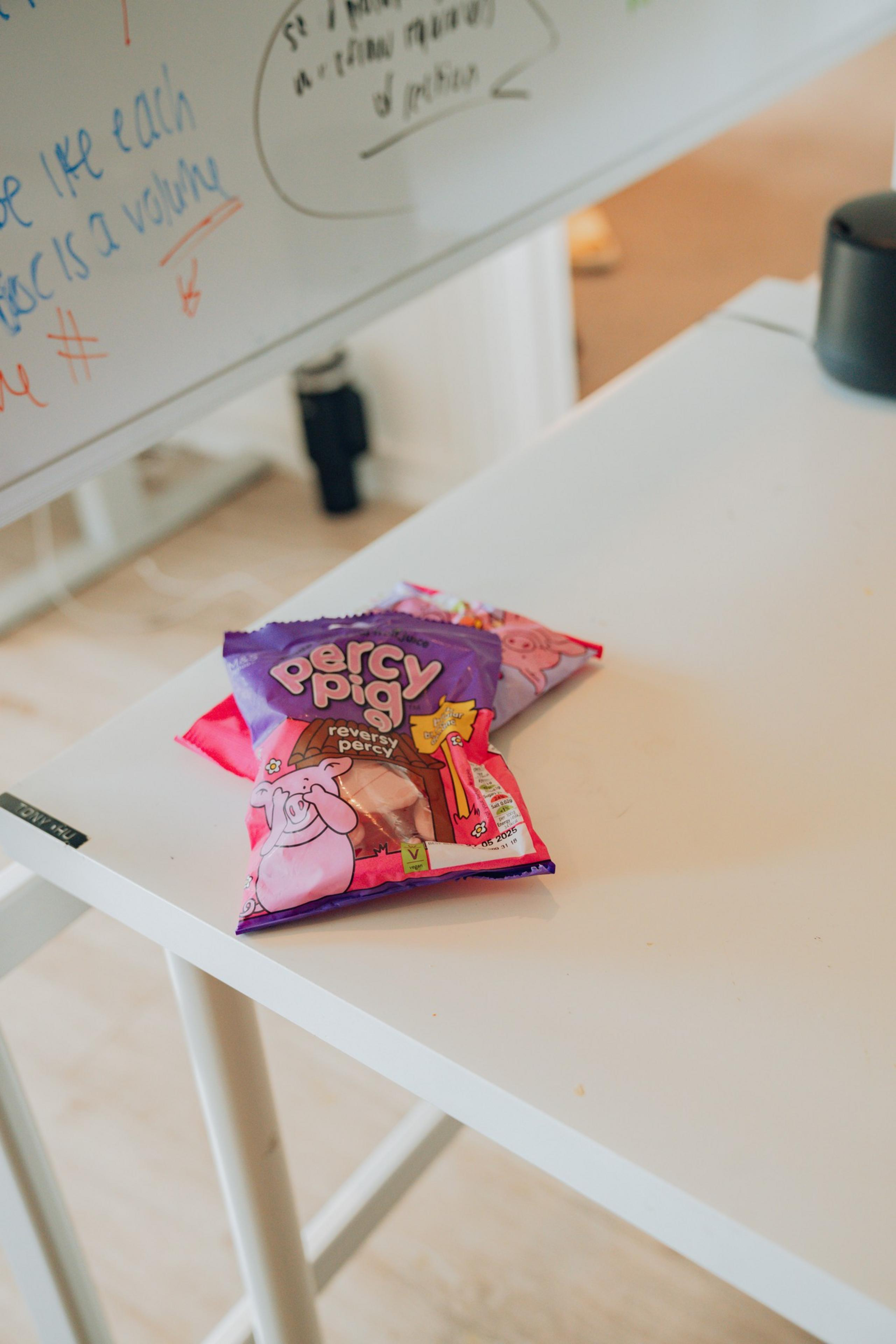 The image shows bags of Percy Pig candy on a white table near a whiteboard with colorful writing. The bags are brightly colored and prominently displayed.