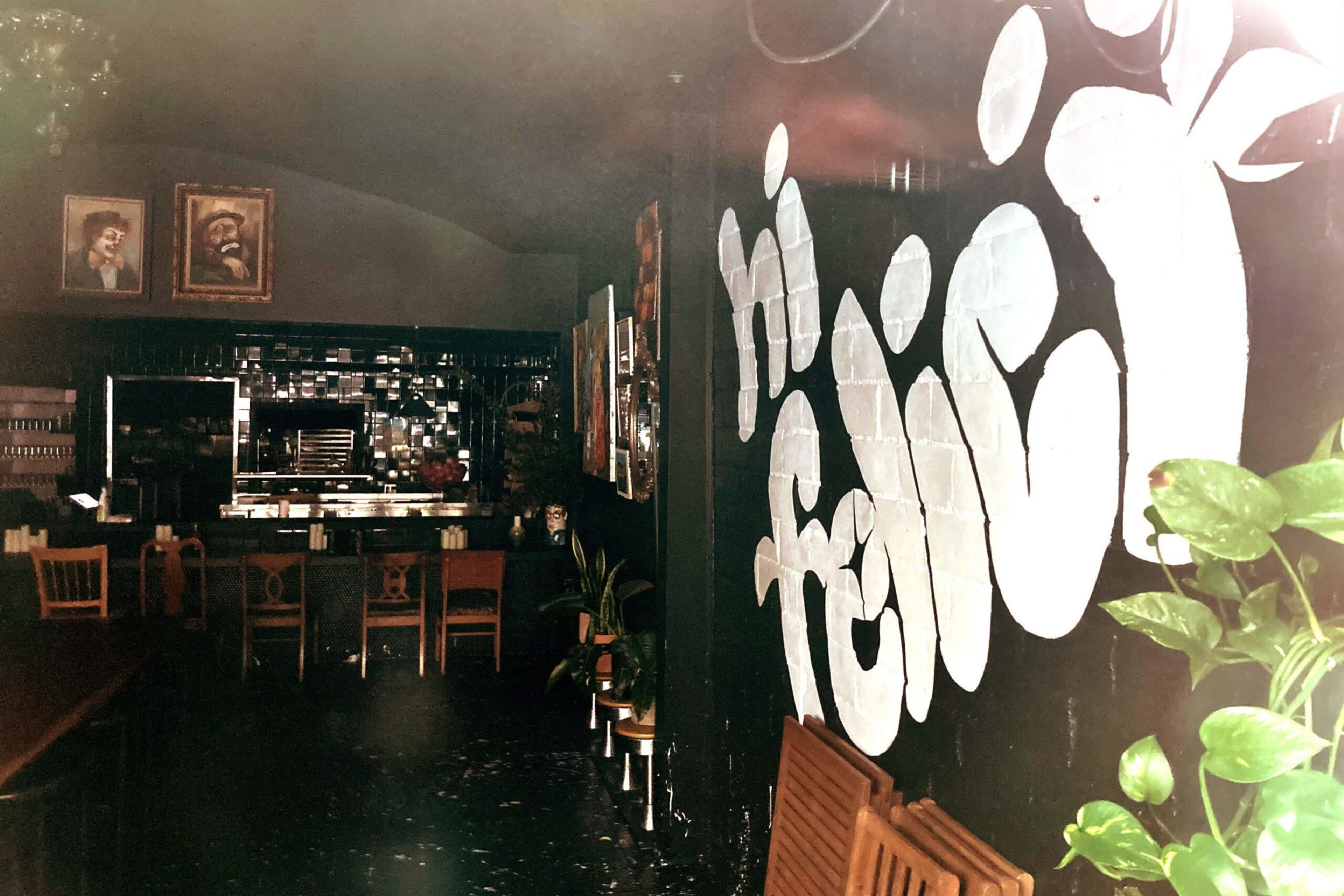 A dark room with wooden chairs around tables, framed portraits on the wall, and a large white graffiti-style "hi friend" on a black wall with some green plants nearby.