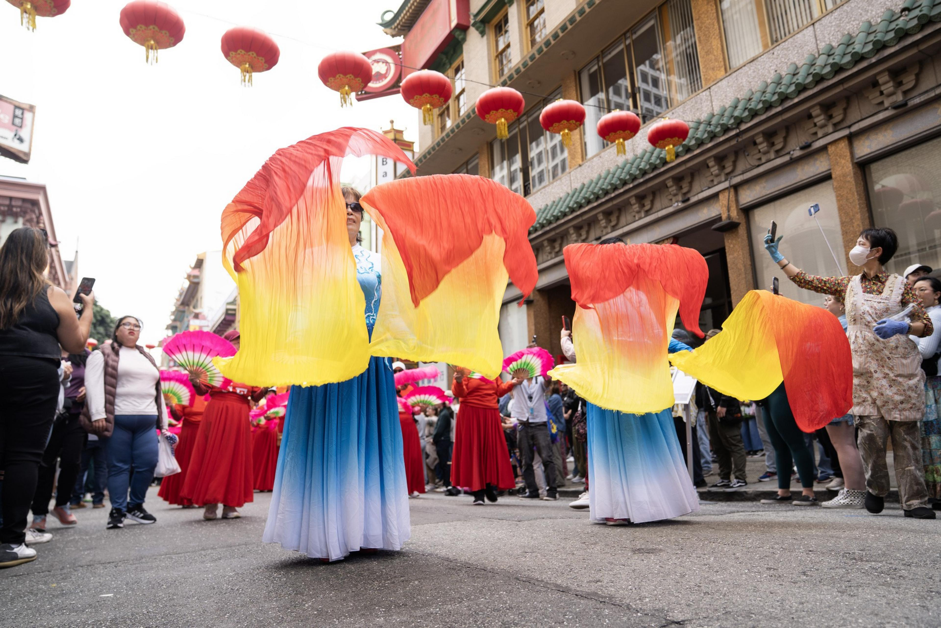 Dancers in blue and white dresses wave large red and yellow silk fans during a street parade with red lanterns hanging overhead and spectators watching.