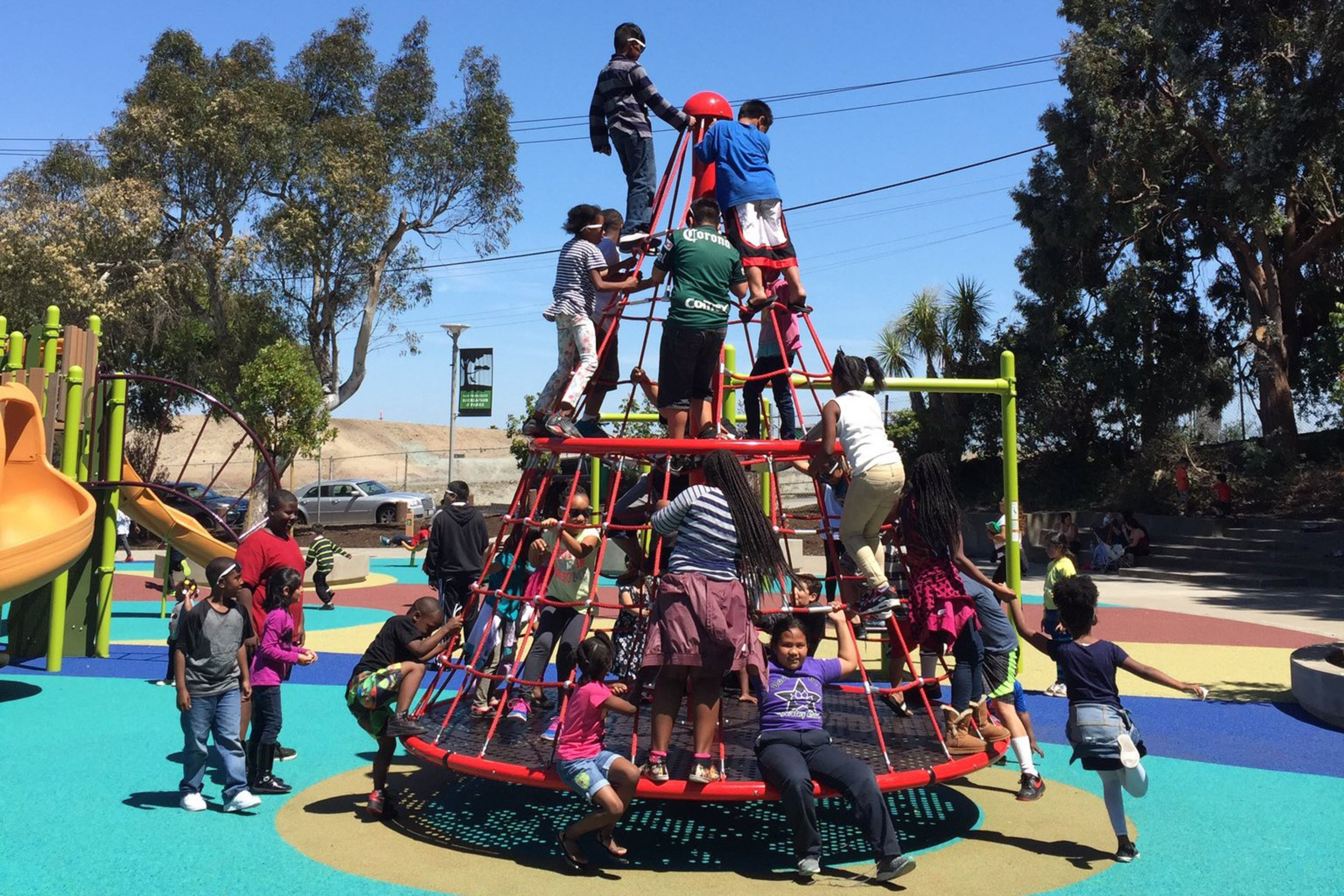 Children of diverse backgrounds play and climb on a red rope pyramid structure in a colorful outdoor playground on a sunny day.