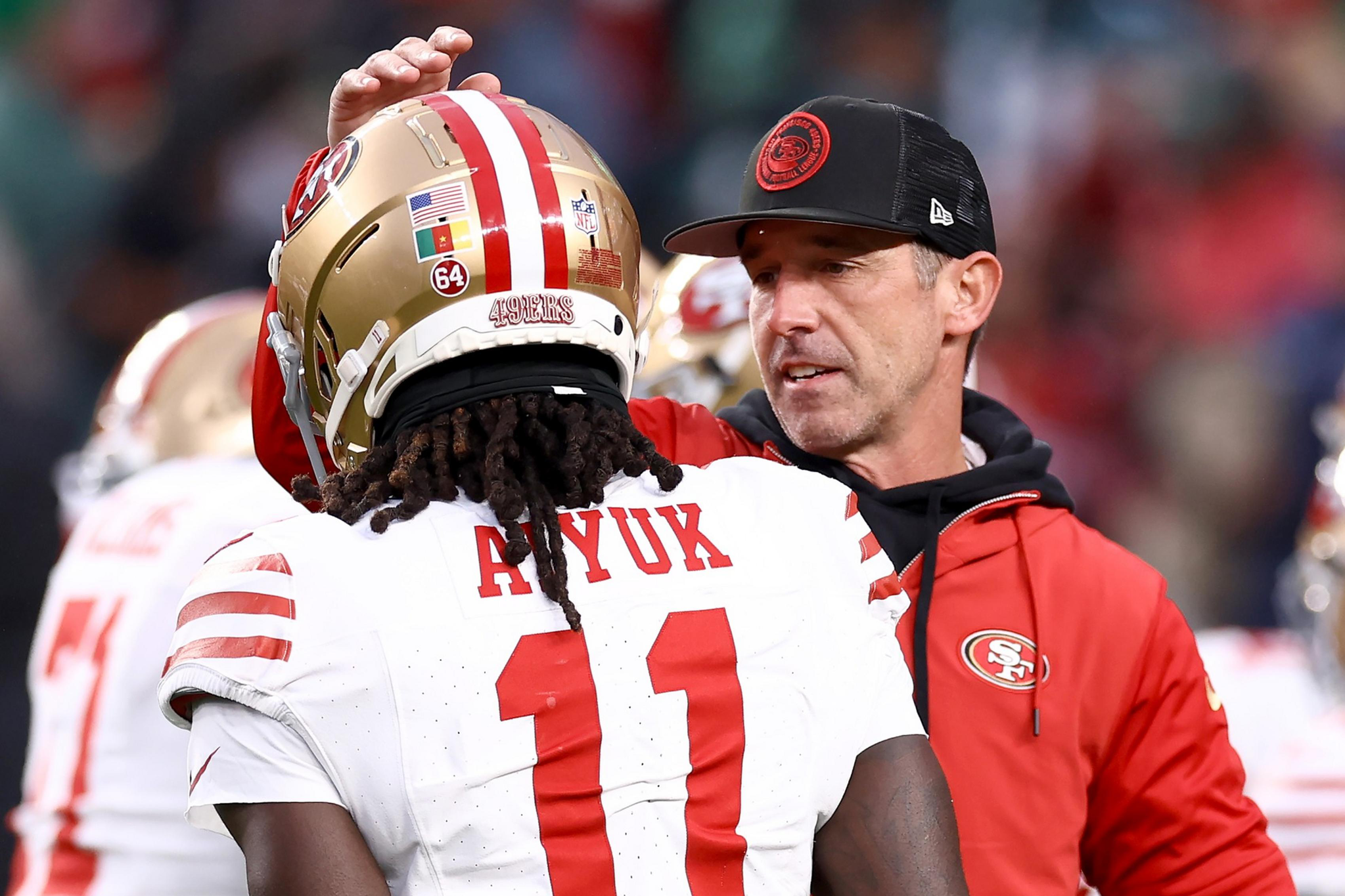 A football coach wearing a red 49ers jacket and black cap talks to a player in a white 49ers jersey with the number 11 and the name Ayuk.