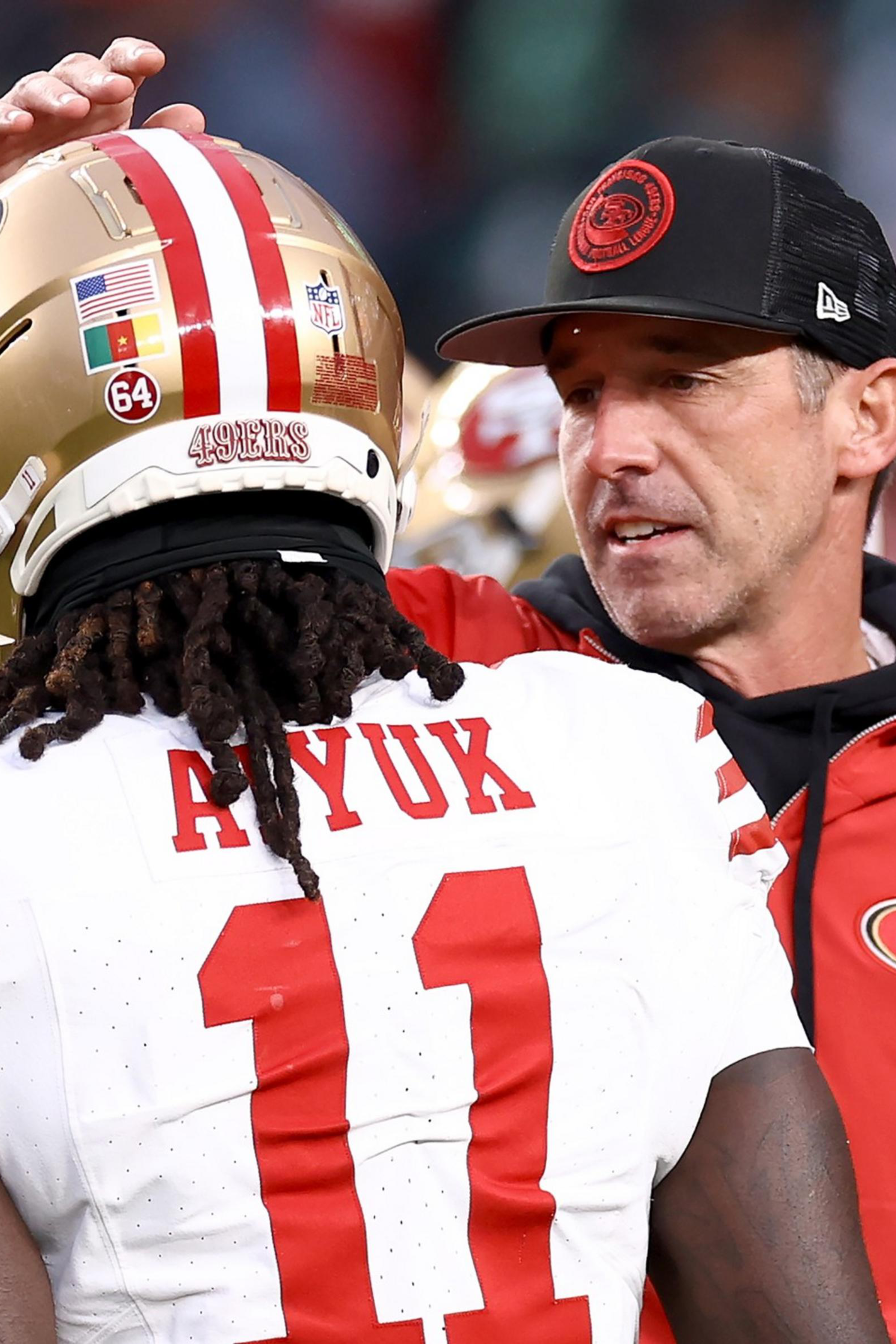 A football coach wearing a red 49ers jacket and black cap talks to a player in a white 49ers jersey with the number 11 and the name Ayuk.