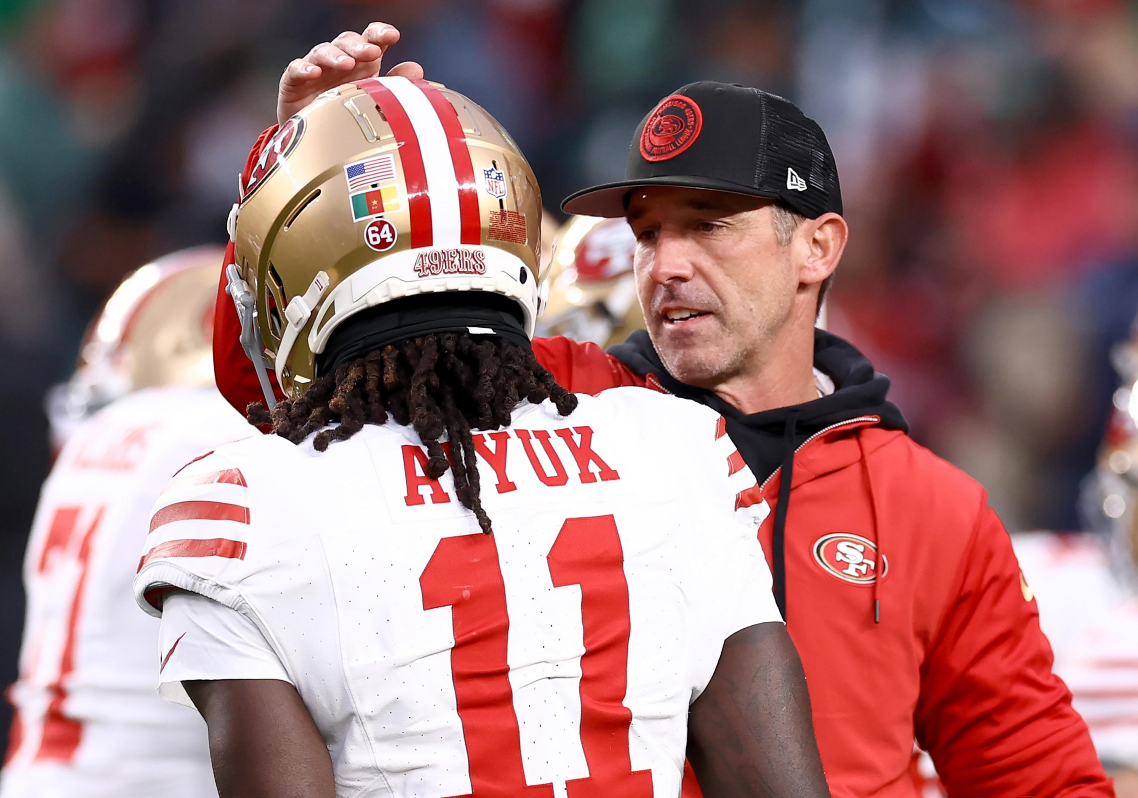A football coach wearing a red 49ers jacket and black cap talks to a player in a white 49ers jersey with the number 11 and the name Ayuk.