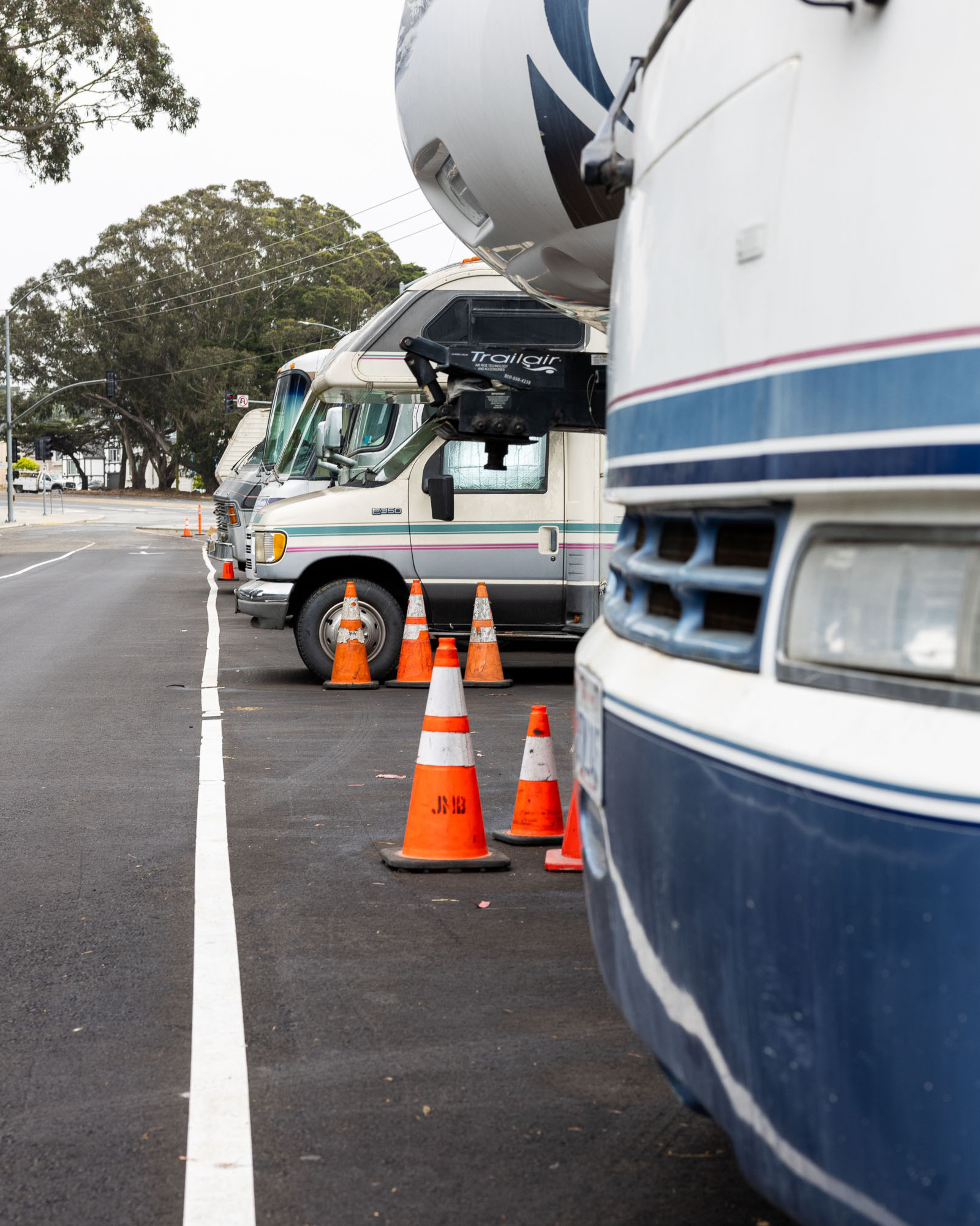 Parked RVs line the side of a street, separated by orange traffic cones. Trees and power lines are visible in the background.