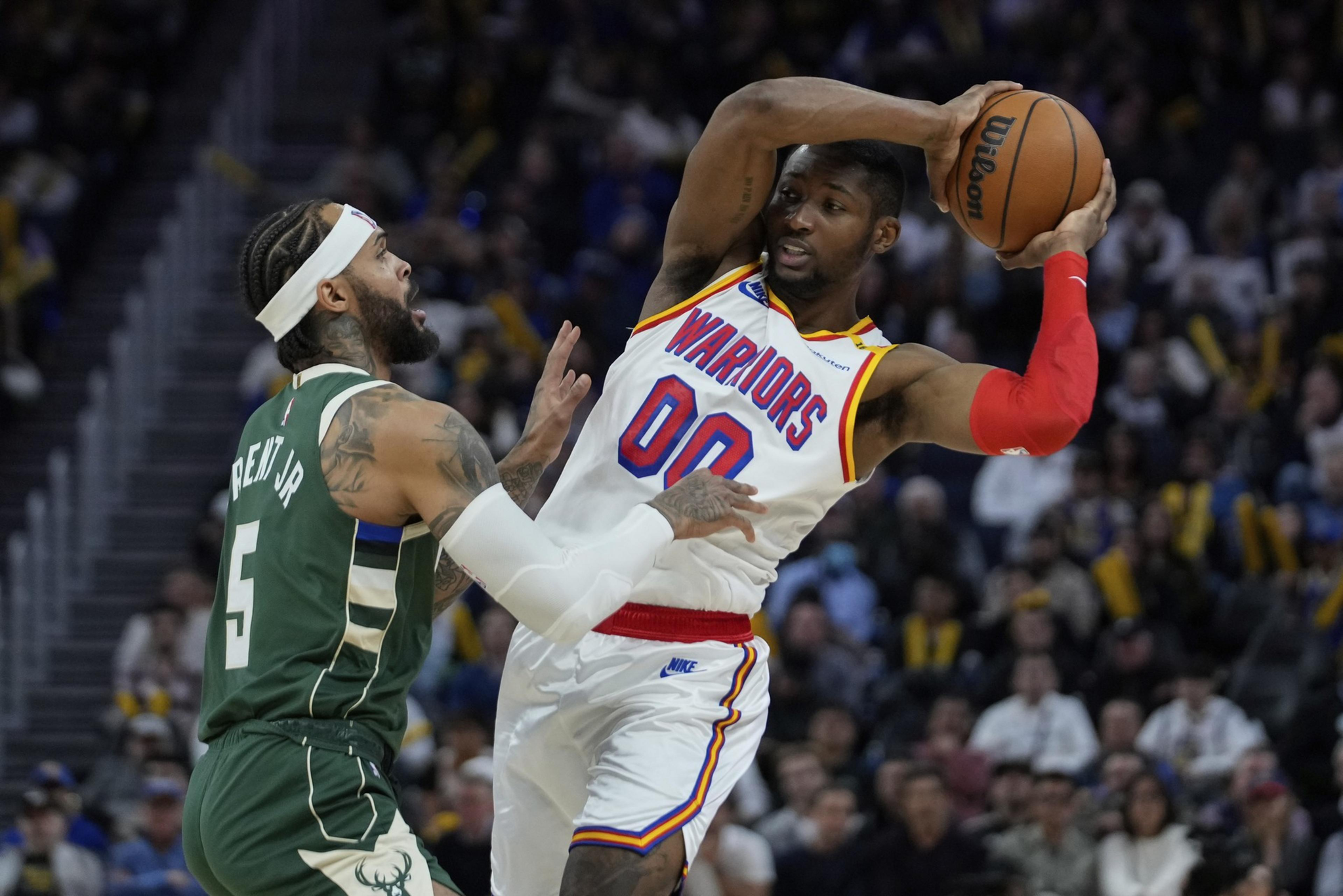 A Warriors player with the ball fends off a defending Bucks player wearing number 5 during an intense basketball game.