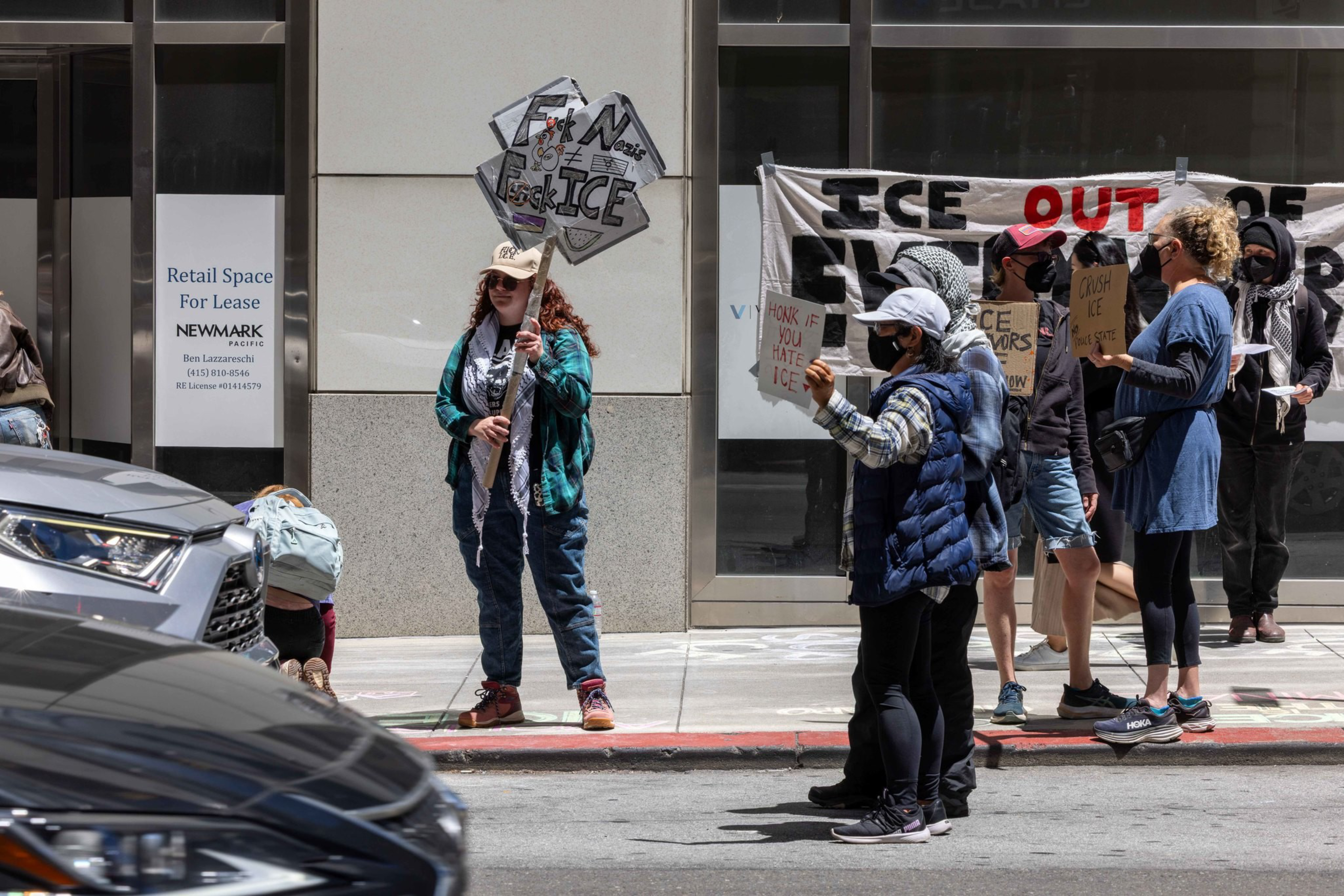 People are protesting on a street, holding signs against ICE. They stand by a building with a &quot;Retail Space For Lease&quot; sign, and cars are visible in the foreground.
