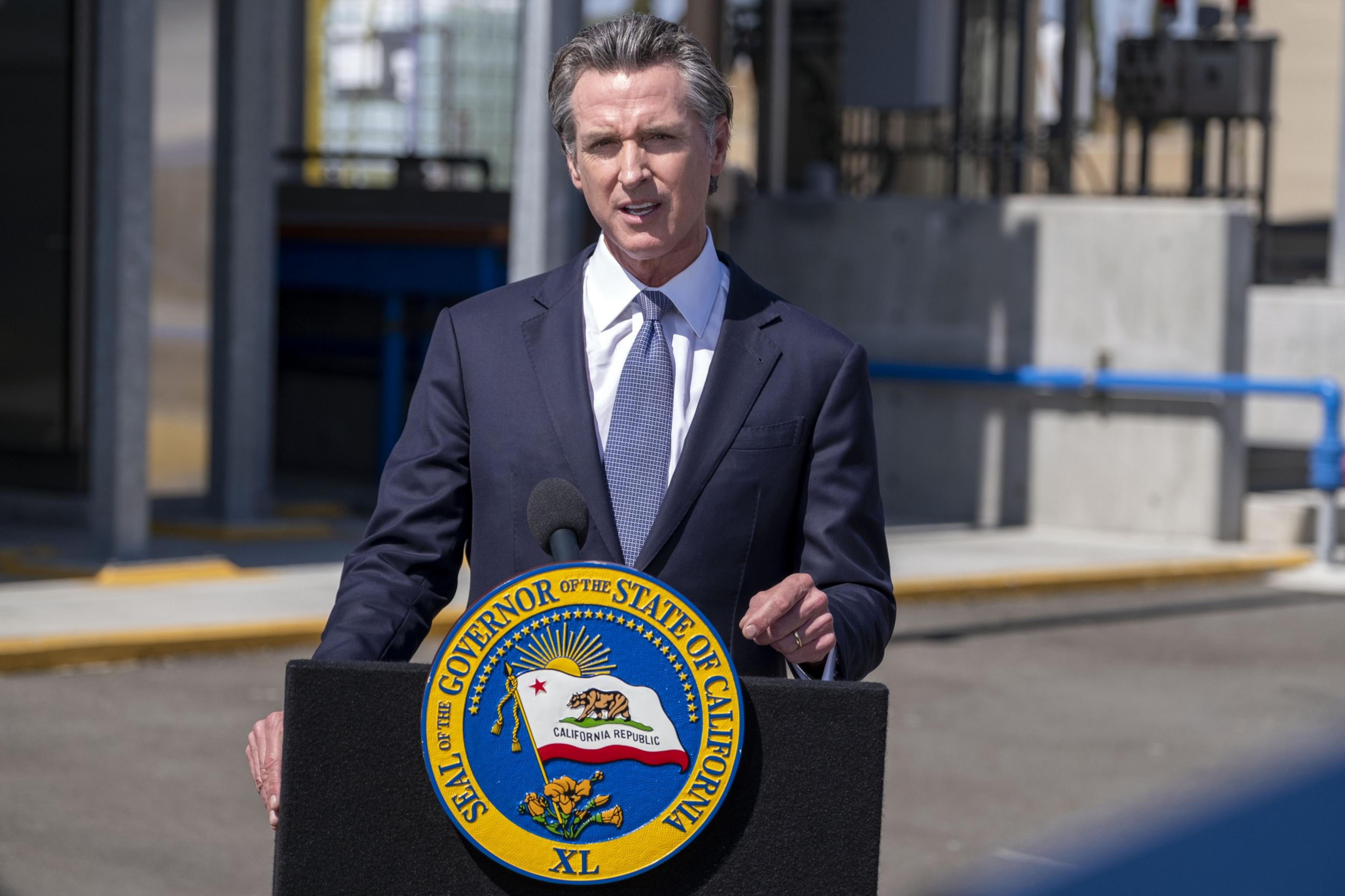 A man in a suit speaks at a podium bearing the California Governor's Seal.