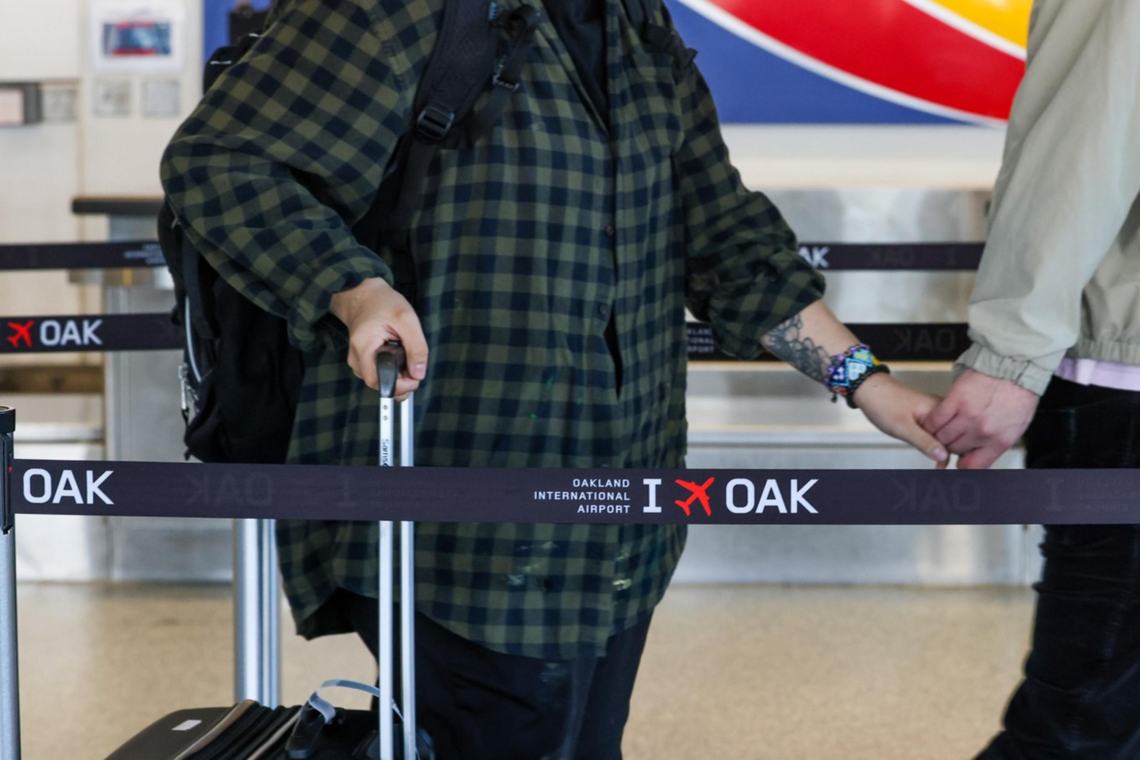 Two individuals at an airport hold hands behind a queue barrier, one with colorful socks, the other with a suitcase.