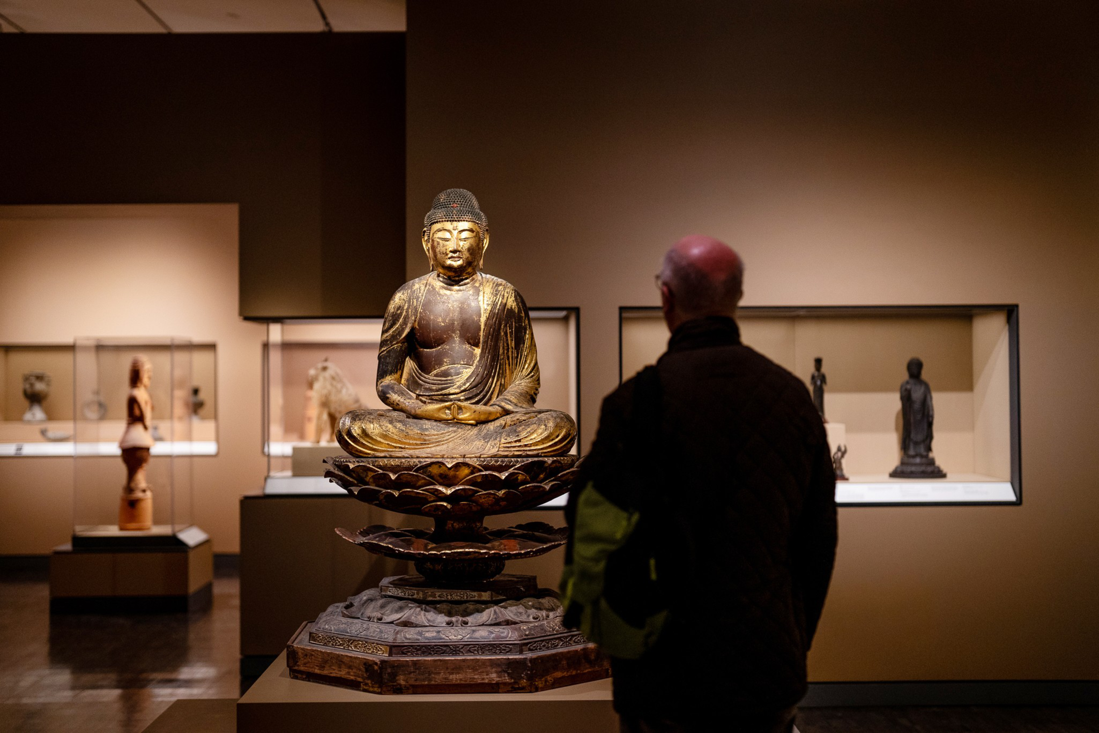 A golden Buddha statue seated on a lotus pedestal is displayed in a museum, while a person with a bald head and glasses observes it closely.