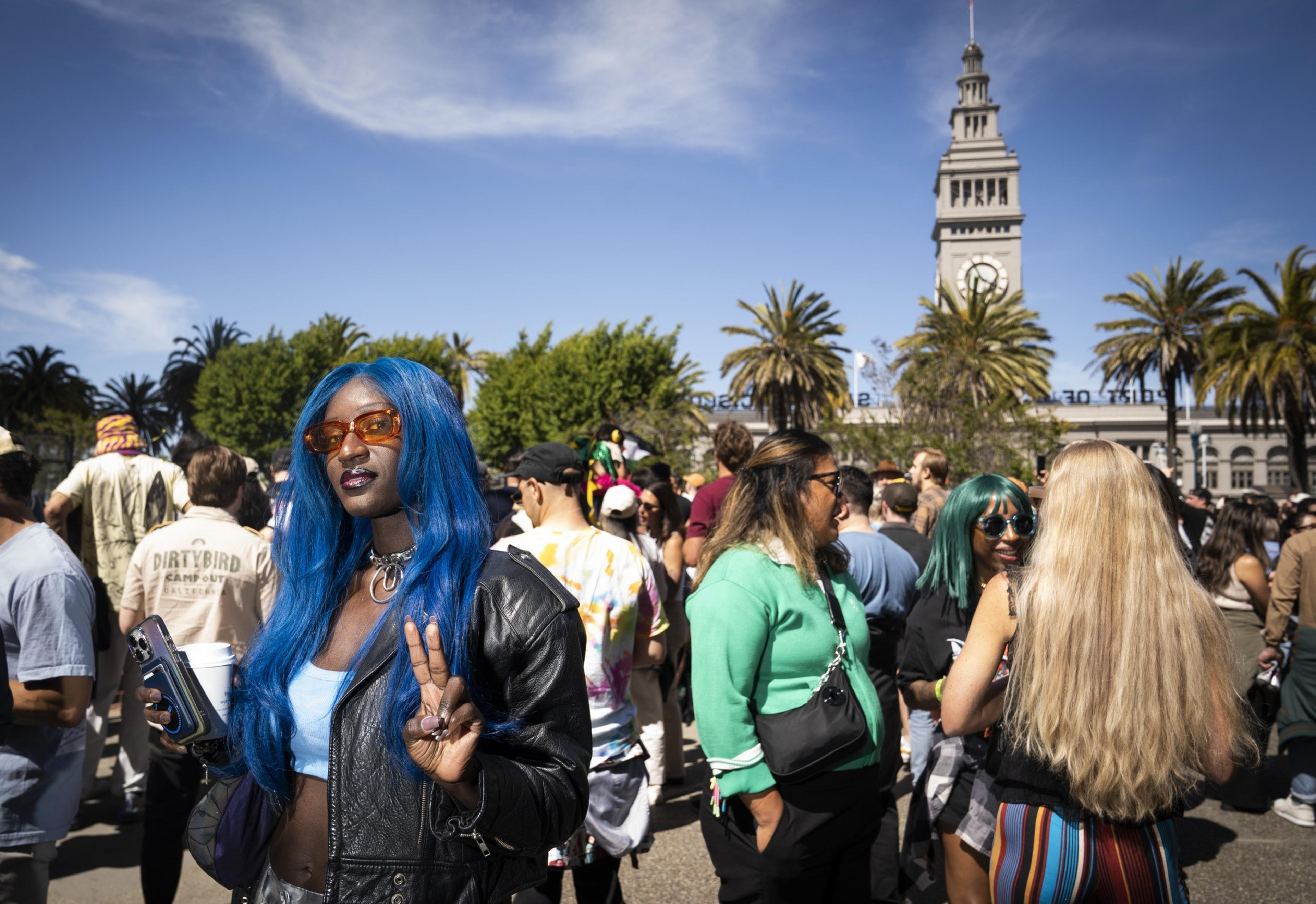 A person with long blue hair and orange sunglasses flashes a peace sign at a crowded outdoor event. A clock tower and palm trees are visible in the background.