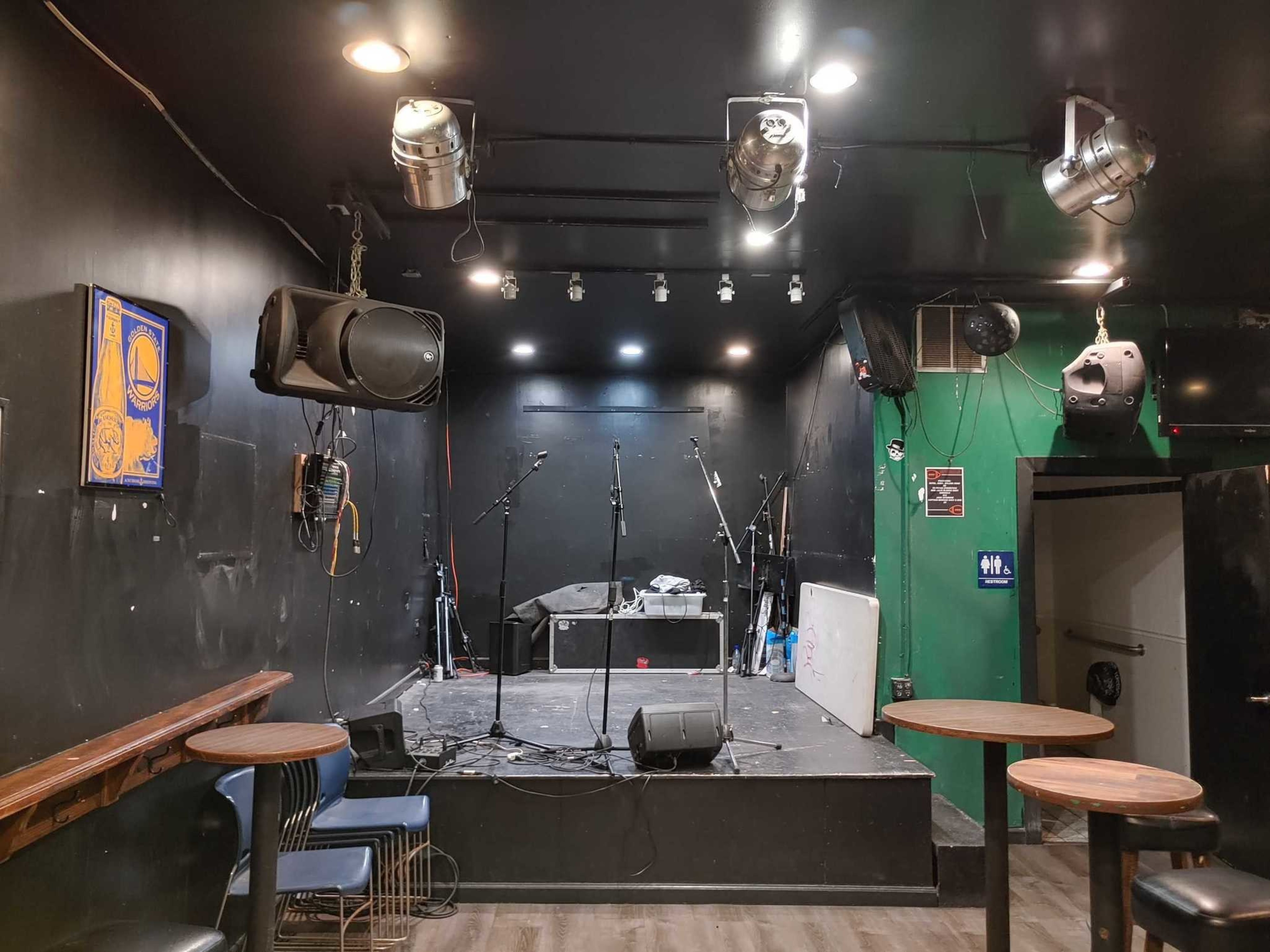 A small, empty stage with microphone stands and speakers is set against dark walls, surrounded by stools and round wooden tables in a cozy room.