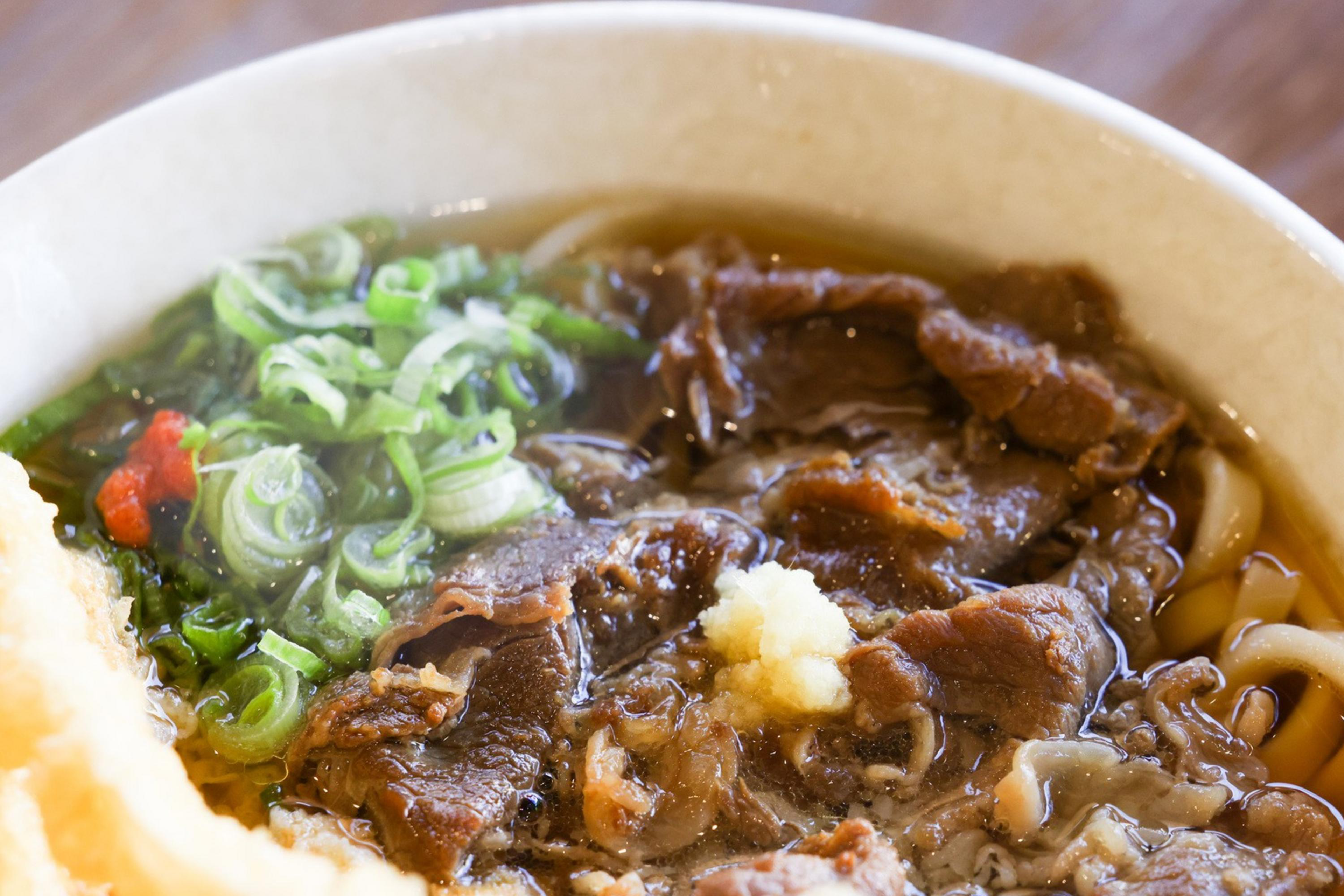 A bowl of beef udon with tempura, garnished with green onions and red spices.