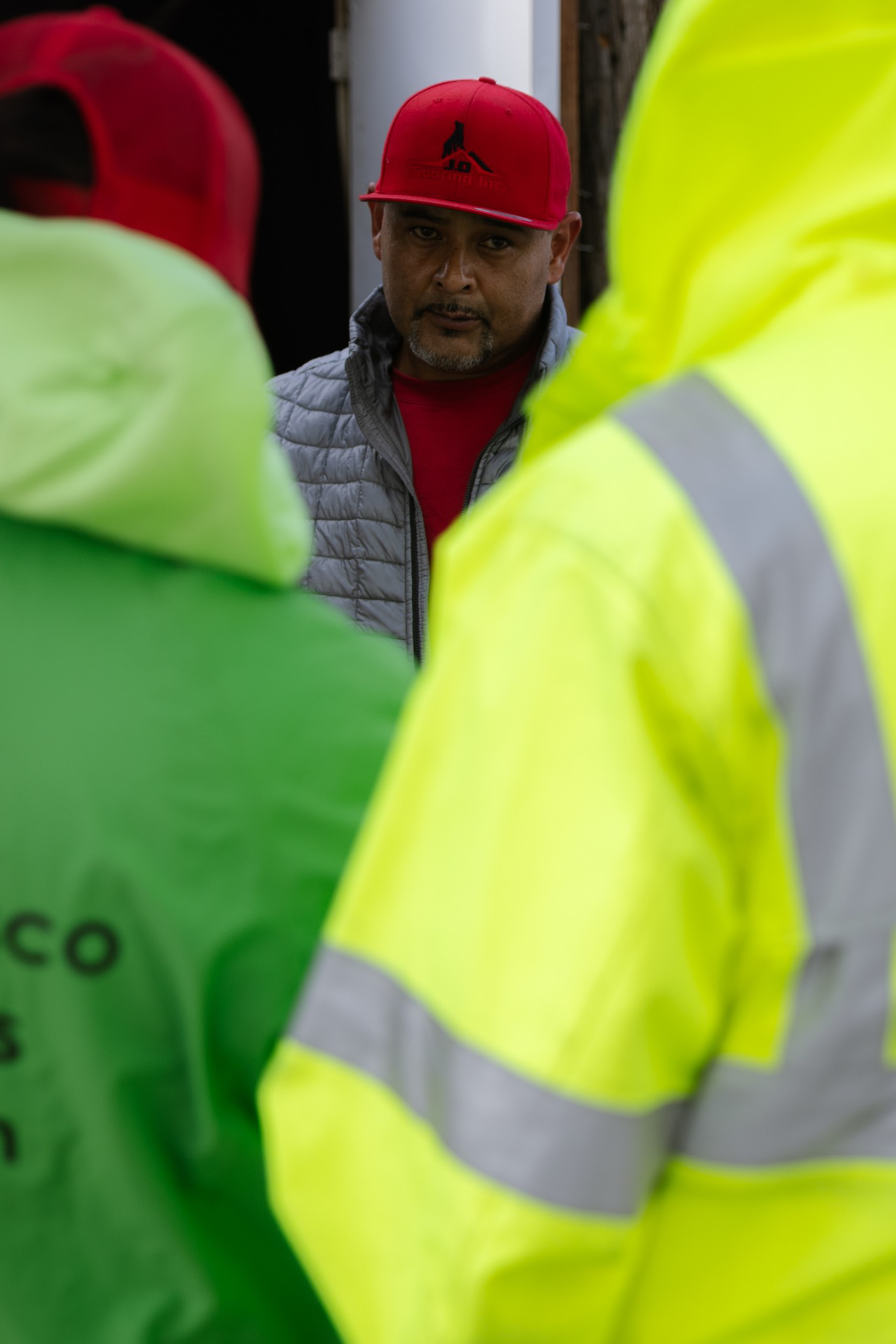 A man in a red cap and gray jacket looks intently ahead, partially framed by two brightly colored high-visibility jackets, one neon green and one yellow.