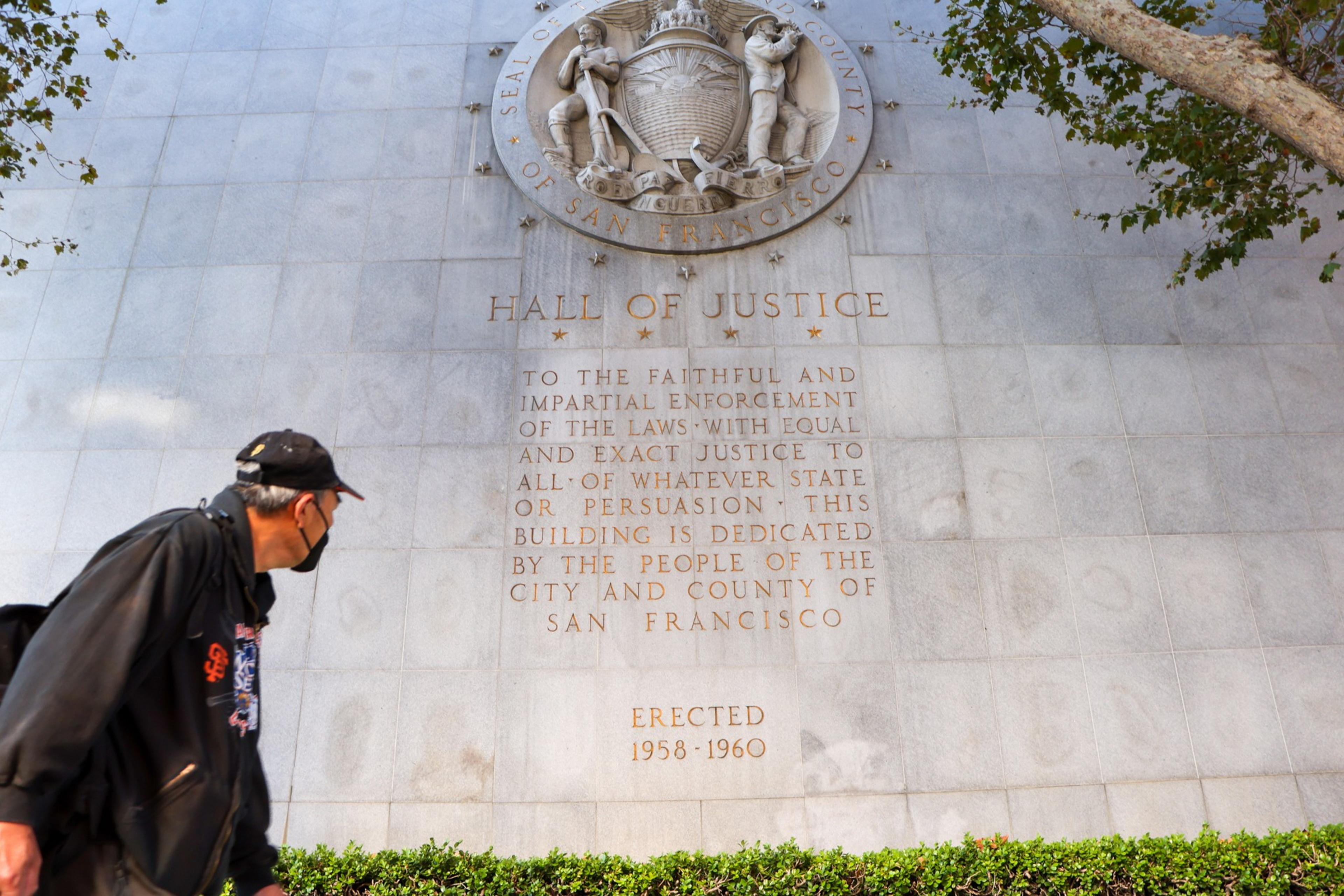 A man in a black jacket and cap looks up at a marble wall engraved with the San Francisco Hall of Justice dedication and the city seal above it.