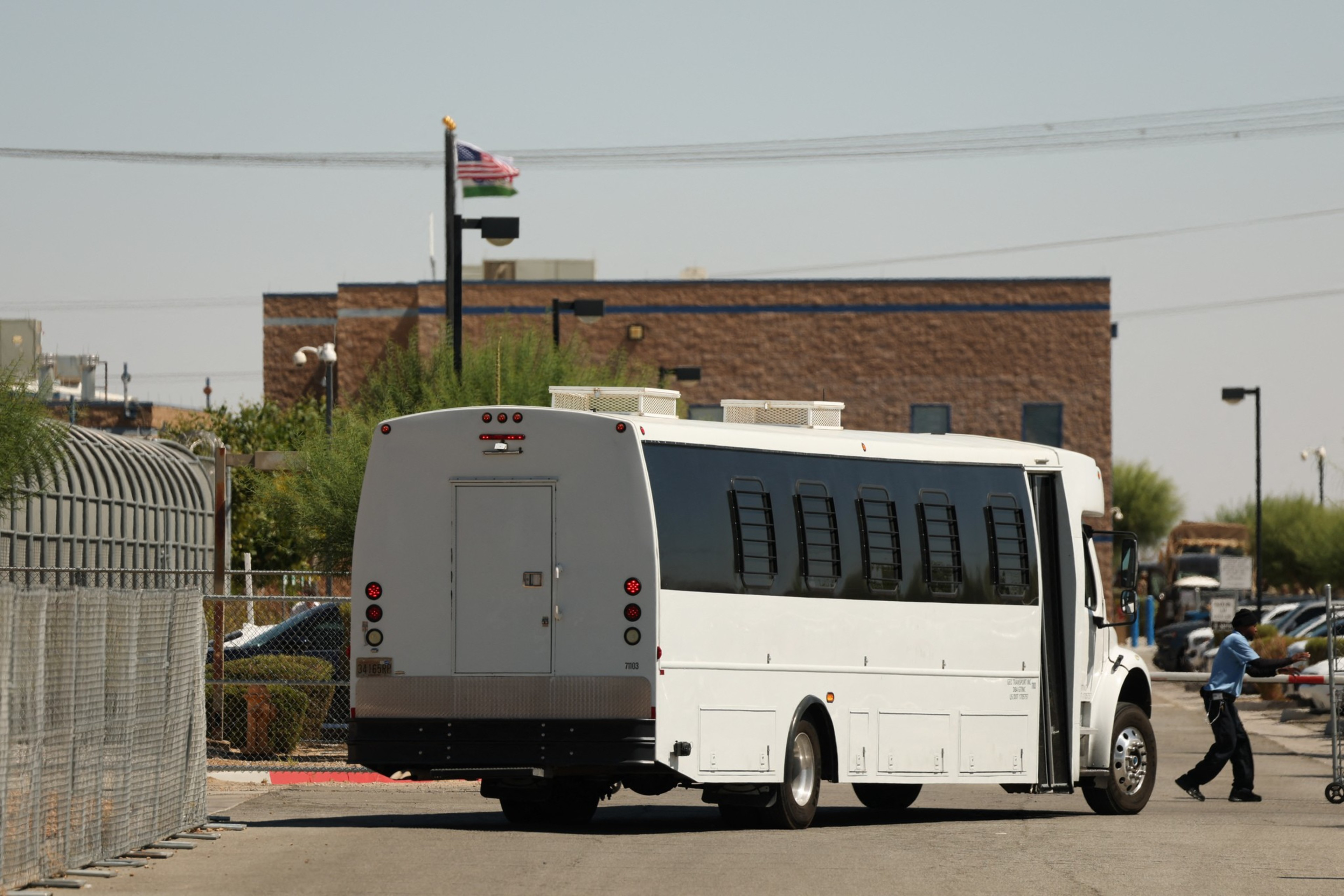 A white bus with barred windows is parked near a chain-link fence. An American flag waves in the background, and a person pushes a cart nearby.