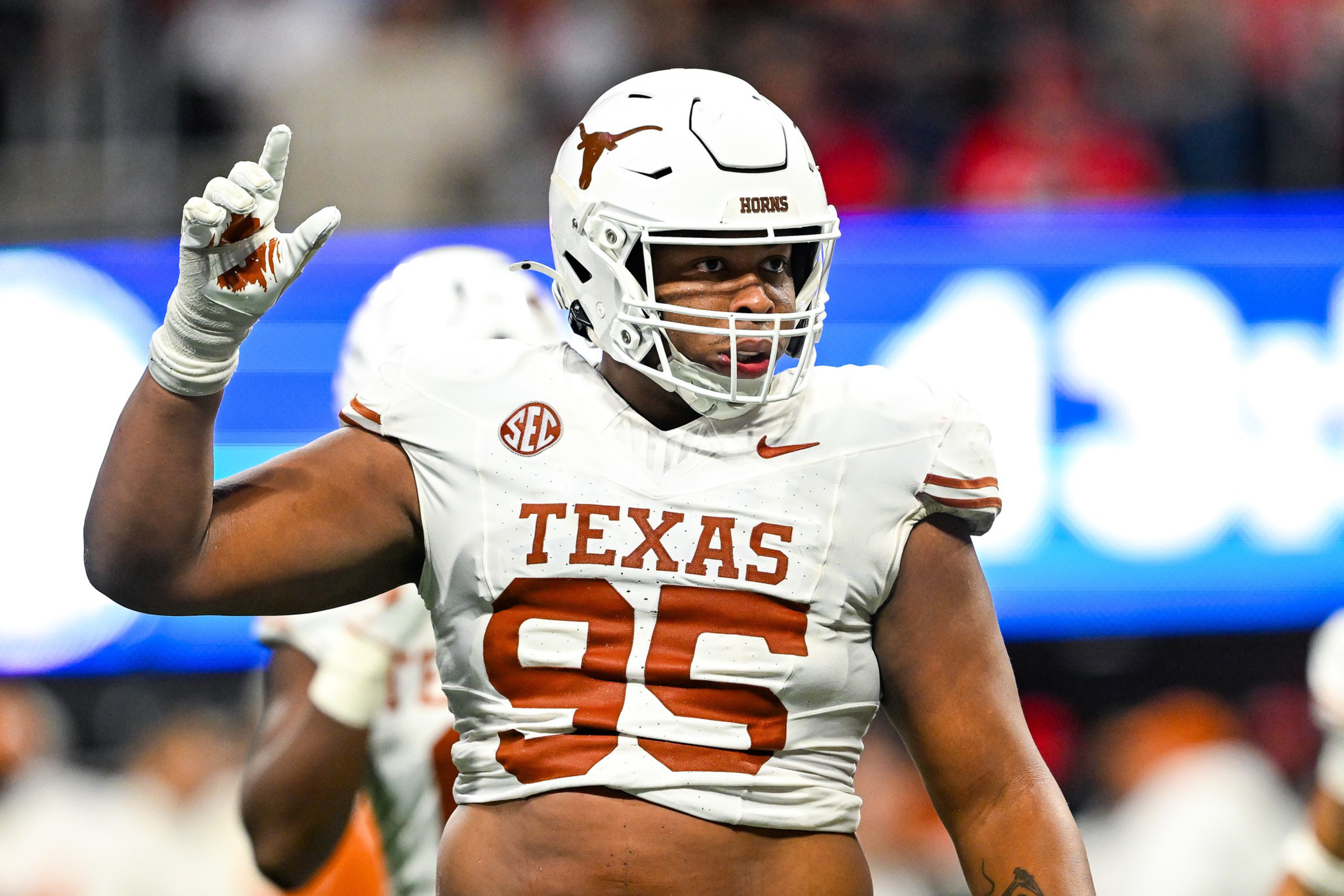 A football player from Texas, wearing a white jersey with the number 95, raises his hand, making the "Hook 'em Horns" sign, during a game.