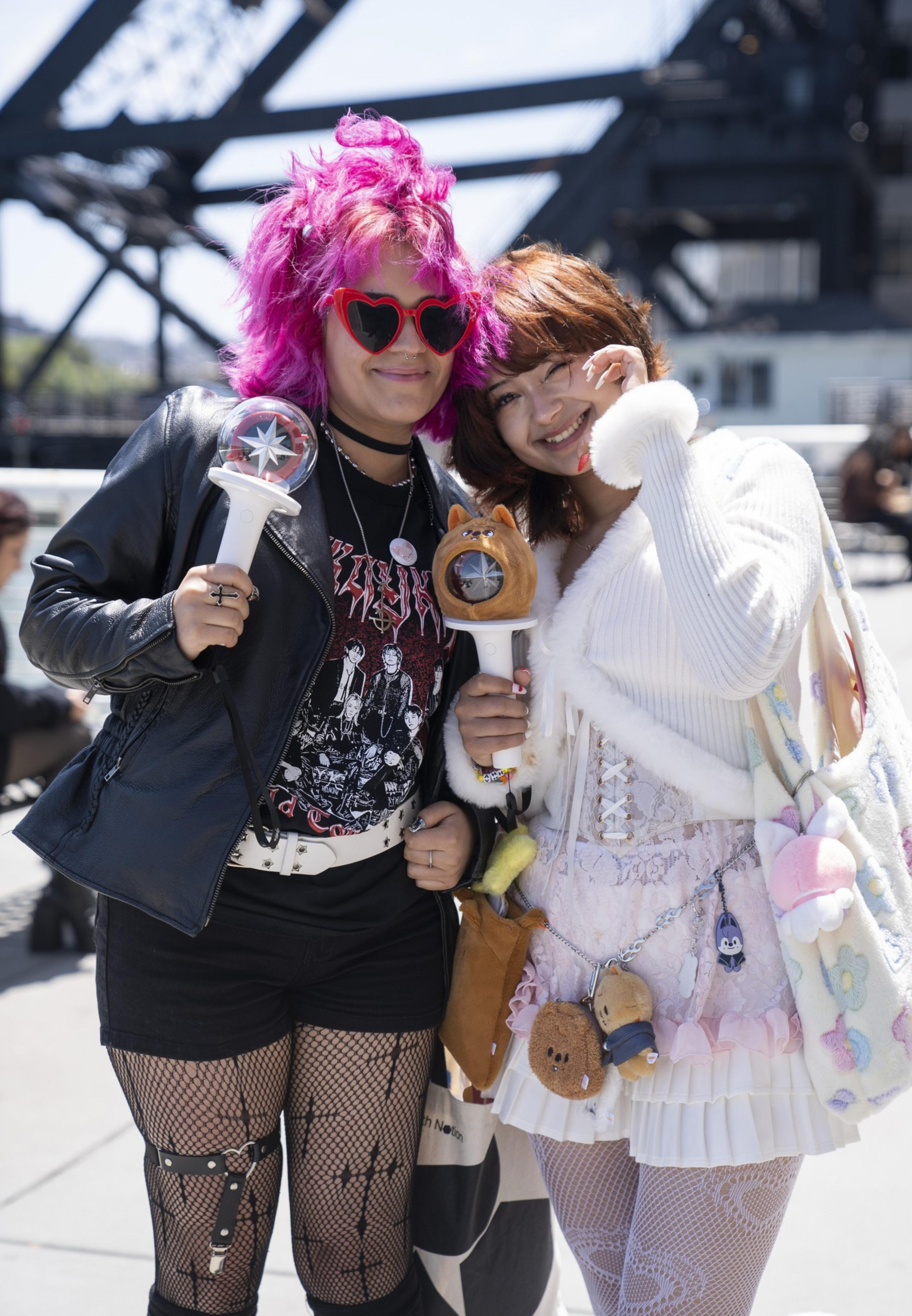 Two friends pose happily outdoors. One has bright pink hair, heart-shaped sunglasses, and a leather jacket. The other wears a fluffy white outfit and lace details.
