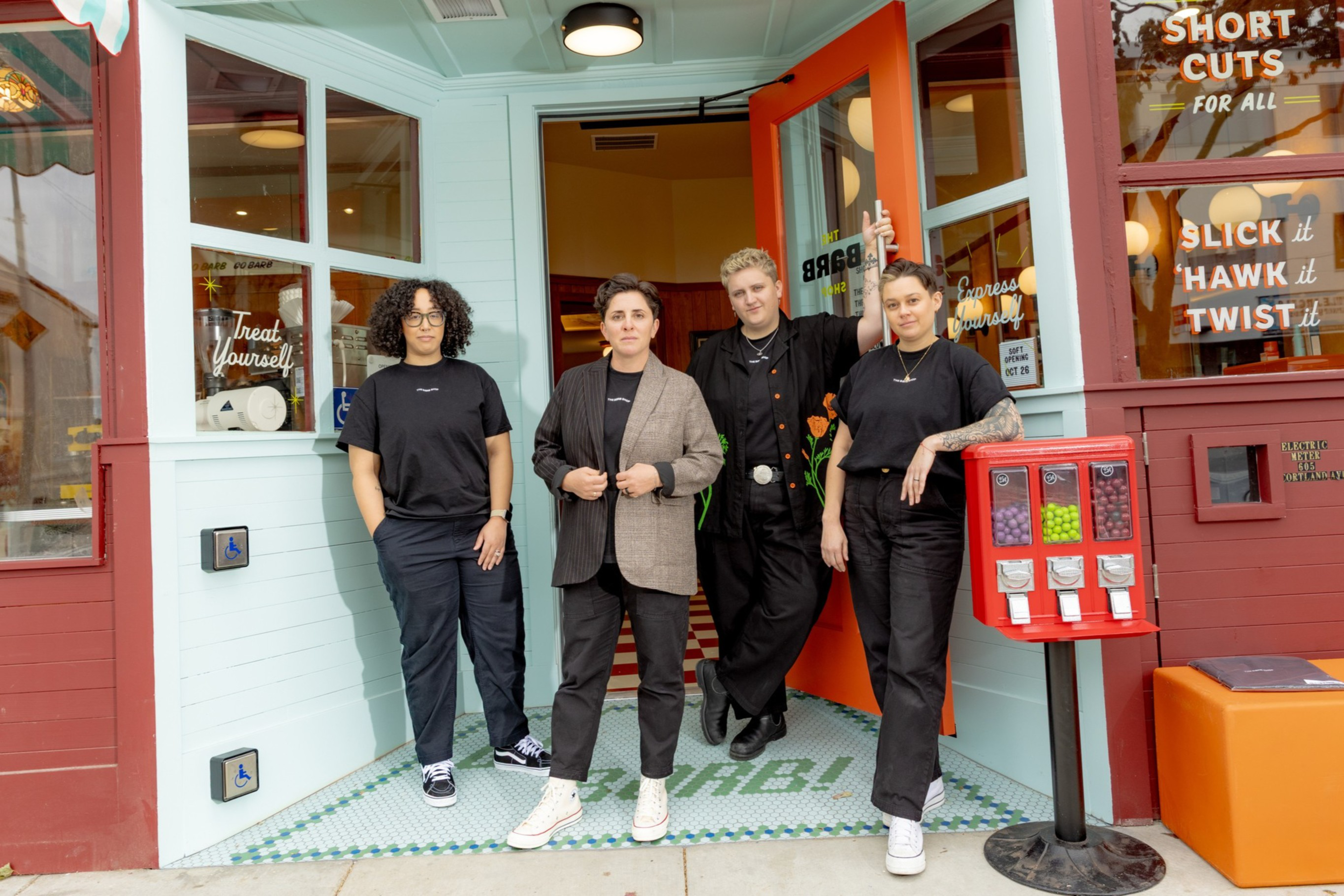 Four people stand casually in front of a retro-style shop with teal walls, an orange door, and a red gumball machine nearby.